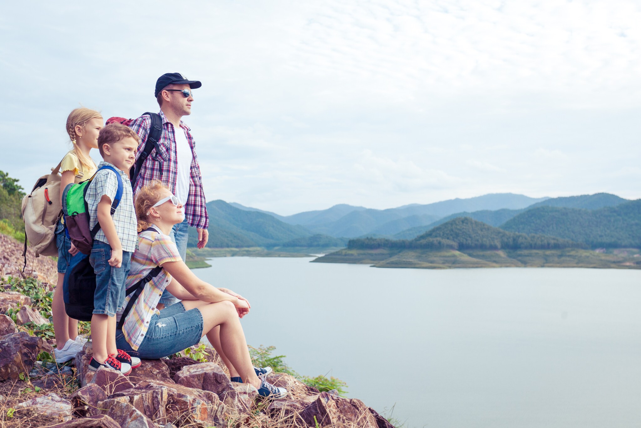 Eine vierköpfige Familie mit Wanderrucksäcken blickt auf einen See, im Hintergrund Berge.