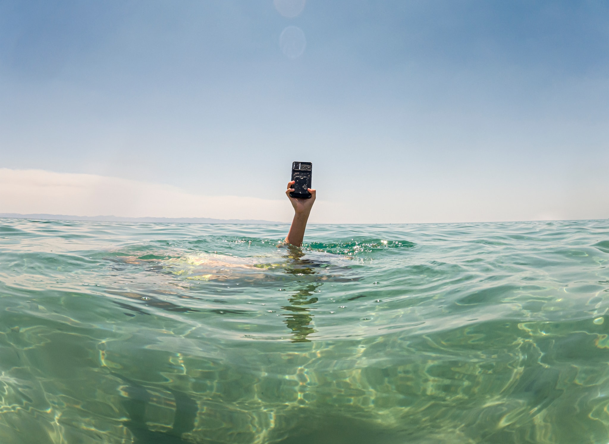 amex-insurance-handy-ins-wasser-gefallen-arm-meer Ein Arm schaut aus dem Meer, in der Hand ein Smartphone.