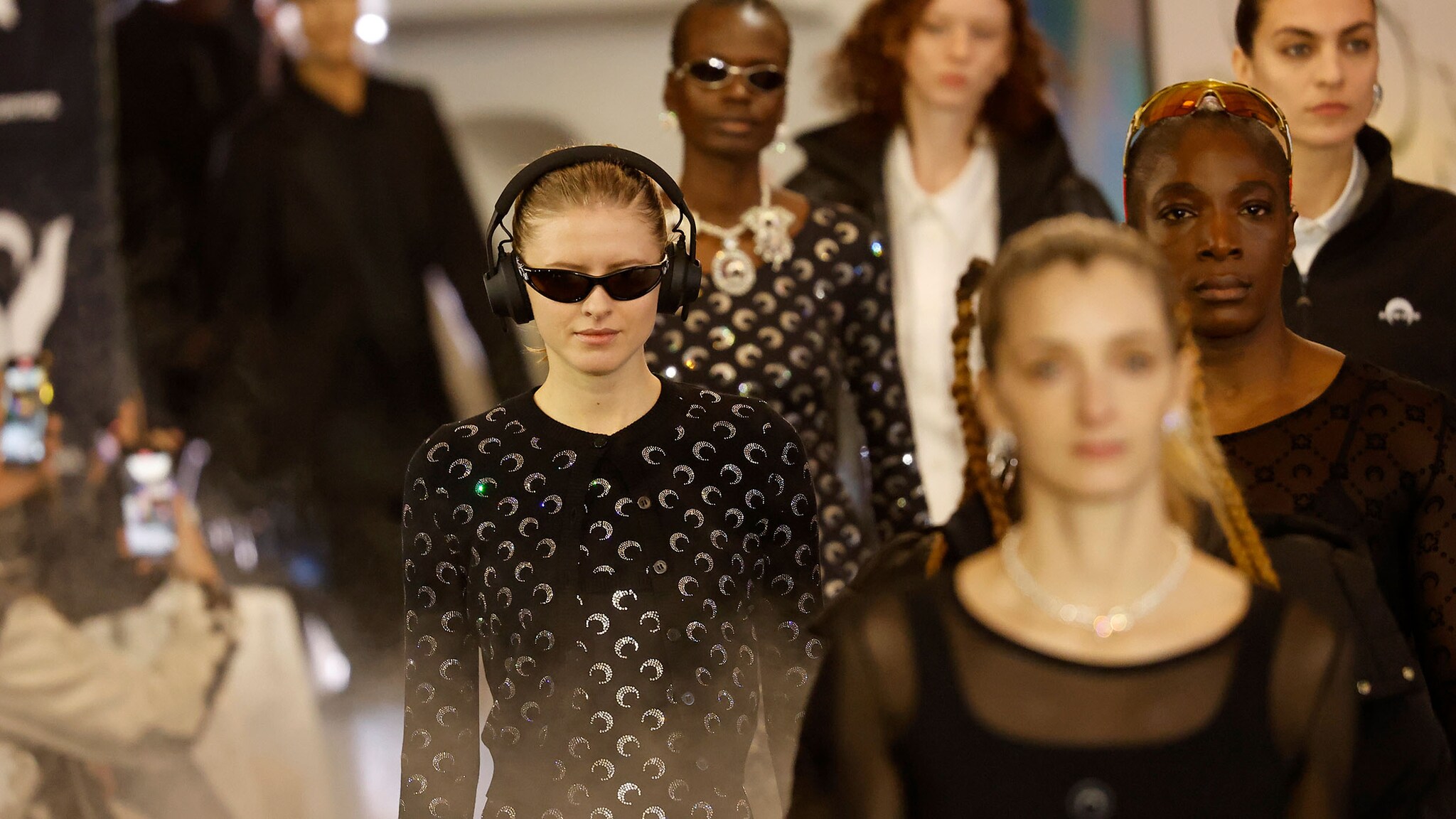 Frontalansicht weiblicher Models in moderner, schwarzer Kleidung und Sonnenbrillen, die über einen Laufsteg laufen bei einer Modenschau.