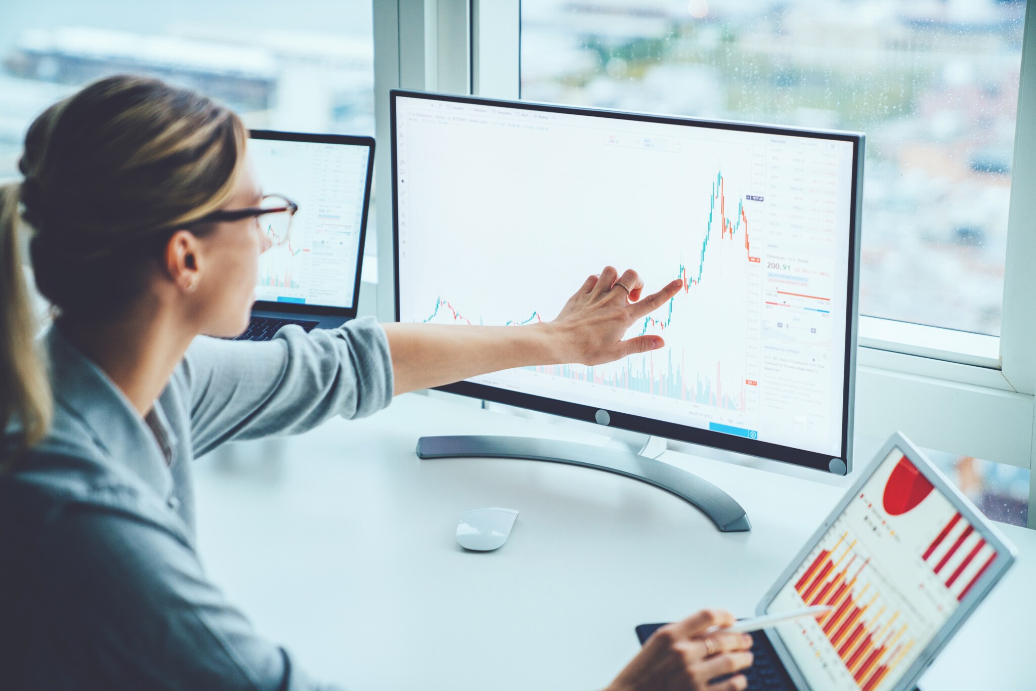 Eine Frau sitzt am Schreibtisch. Mit ihrem Zeigefinger zeigt sie auf auf ein statistisches Diagramm auf dem Computerbildschirm vor ihr.