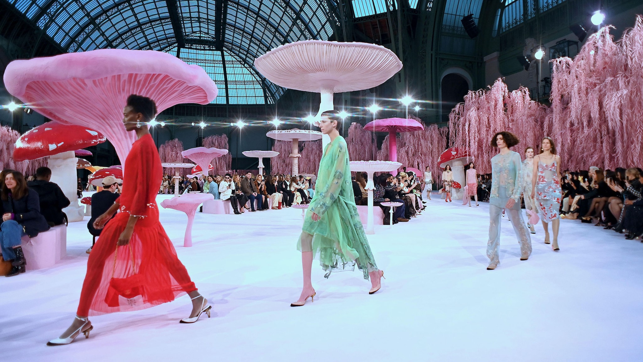 Weibliche Models laufen in halbtransparenter Sommerkleidung über einen Laufsteg mit pinken Pilz-Skulpturen in einer Halle während einer Modenschau mit Publikum.