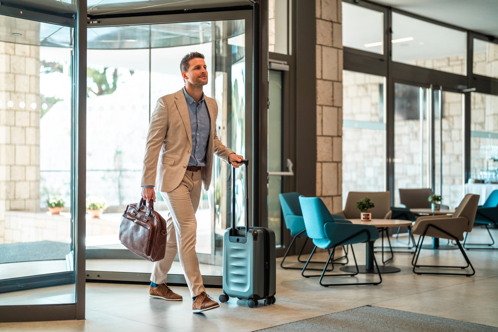 Ein Geschäftsmann mittleren Alters betritt die Lobby eines modernen Hotels, eine braune Ledertasche in der Hand und einen Rollkoffer hinter sich herziehend.