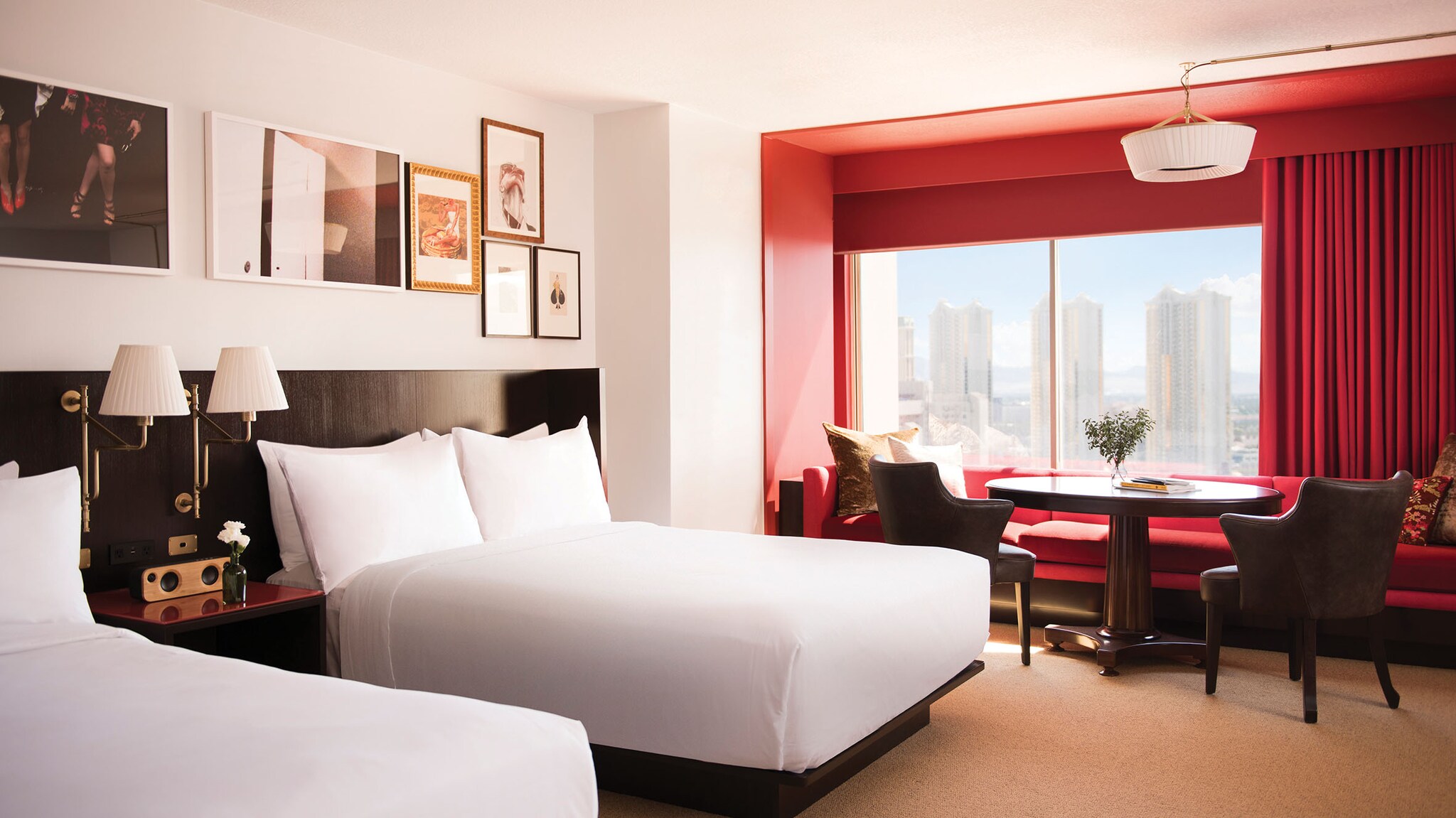 Hotelzimmer mit zwei Doppelbetten vor einer Wand mehreren Bilderrahmen sowie Fensterblick auf eine Skyline mit Hochhäusern. 
