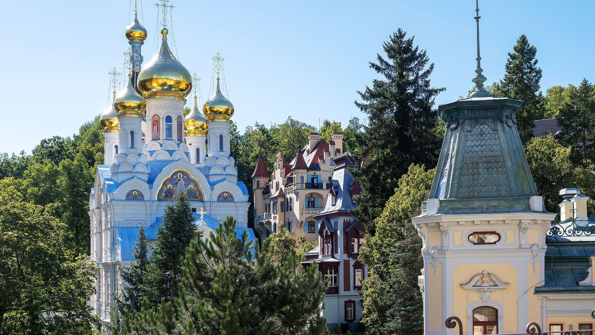 Obere Hälfte einer hellblauen russisch-orthodoxen Kirche mit goldenen Kuppeln neben zwei Villen zwischen grünen Baumwipfeln.