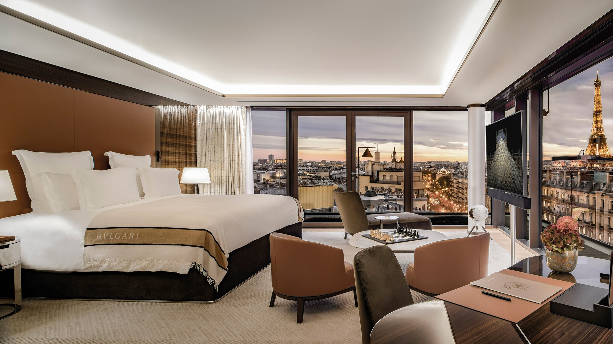 Luxuriöse Hotelsuite mit Blick auf die Skyline von Paris inklusive Eiffelturm durch bodentiefe Panoramafenster am Abend.