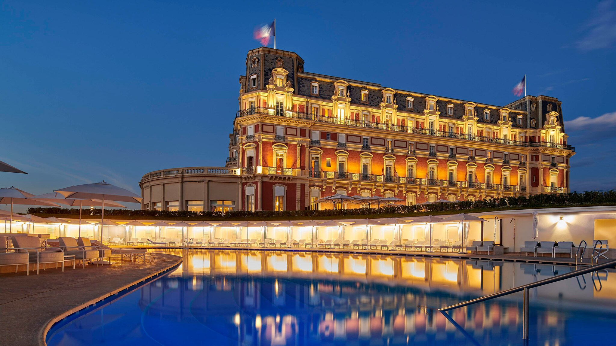 Beleuchtetes Grandhotel mit wehenden französischen Flaggen hinter einem Außenpool mit Cabanas bei Dunkelheit.
