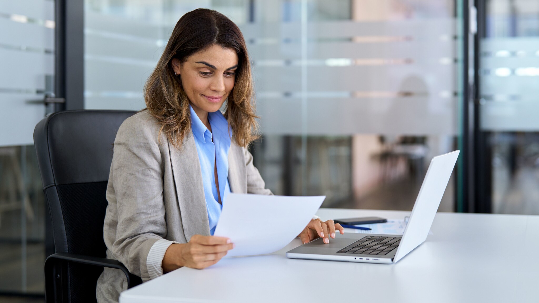 Eine Frau in Business-Kleidung sitzt an einem Schreibtisch und schaut auf ein Blatt Papier in einer Hand, die andere Hand hat sie auf dem Laptop-Touchfeld.