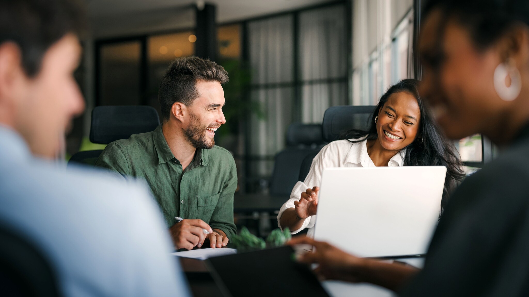 Vier Personen sitzen lachend an einem Schreibtisch mit Laptop und Arbeitsmaterialien in einem Büro.