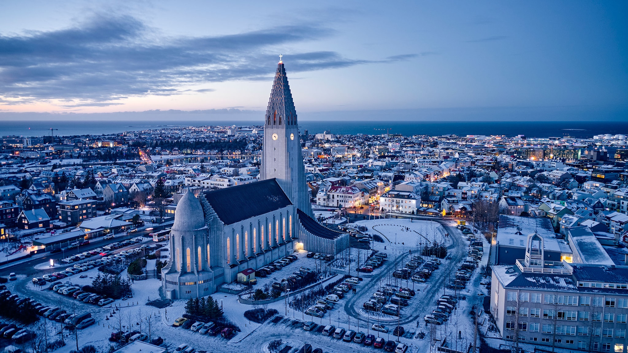 Luftaufnahme von schneebedecktem Stadtpanorama von Reykjavík mit Kirche am Meer im blauen Licht bei Dämmerung.
