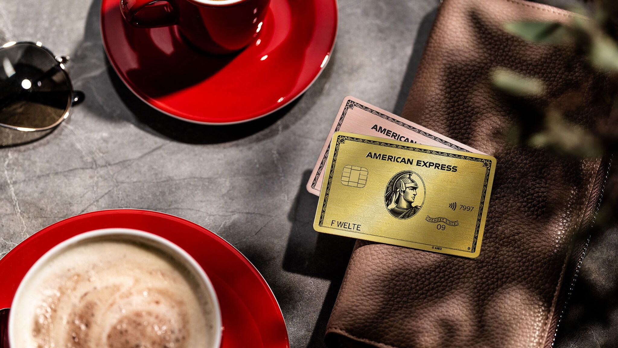 Eine goldene und eine rosafarbene Kreditkarte von American Express liegen übereinander auf einer Geldbörse neben zwei roten Cappuccinotassen auf einem Marmortisch.