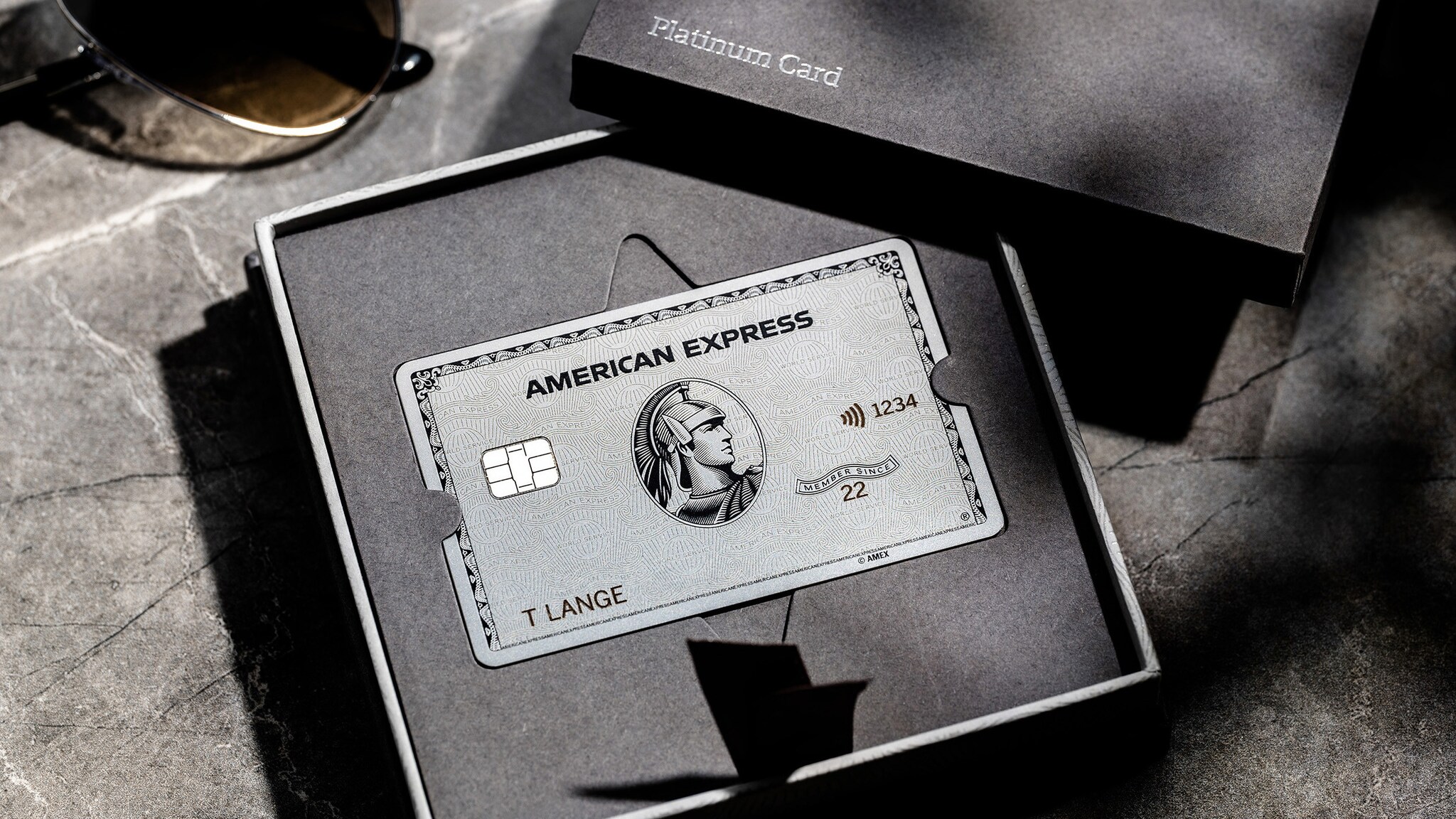 Eine silberne Platinum Card von American Express in einer Präsentationsbox aus grauer Kartonage auf einem grauen Marmortisch.