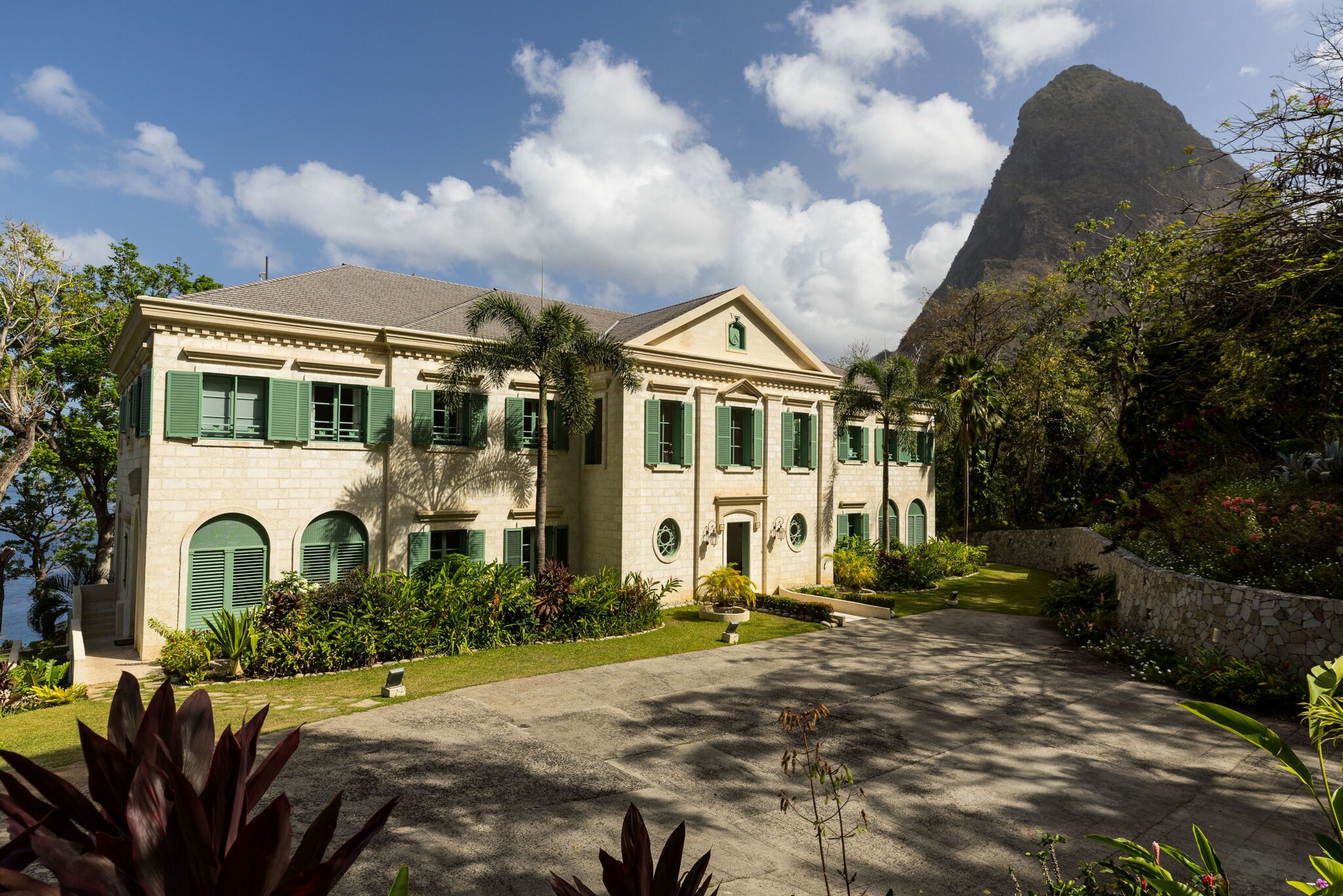 Helle Villa mit grünen Fensterläden in einem tropischen Garten auf St. Lucia. Im Hintergrund erhebt sich ein steiler, bewaldeter Piton unter blauem Himmel mit weißen Wolken.