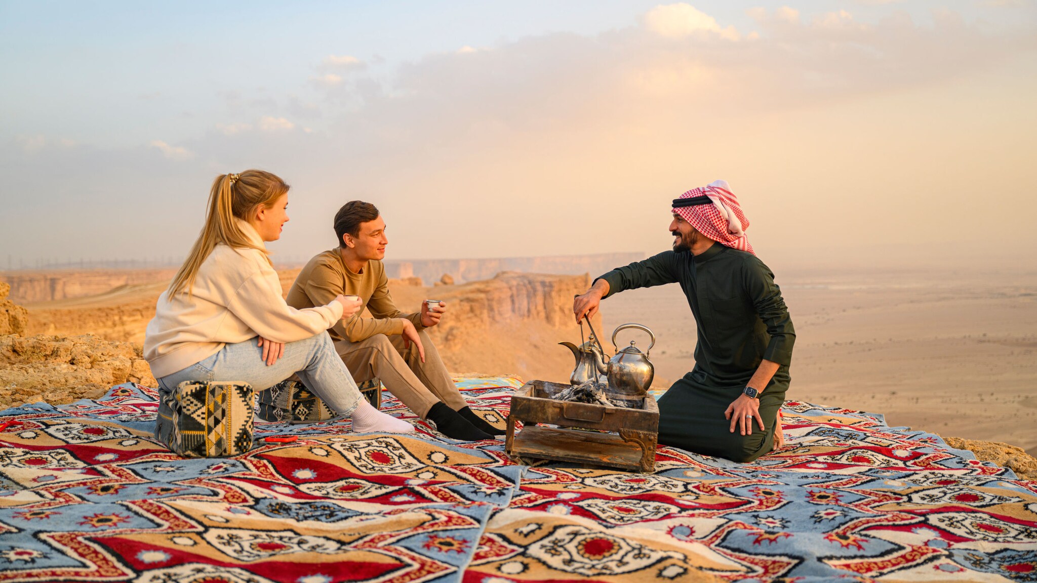 Ein Mann und eine Frau sitzen mit Teetassen in den Händen auf einem Orientteppich an einer Klippe in einer Wüstenlandschaft, ihnen gegenüber kniet ein Mann mit rot-weiß kariertem Kopftuch vor einer Teekanne.