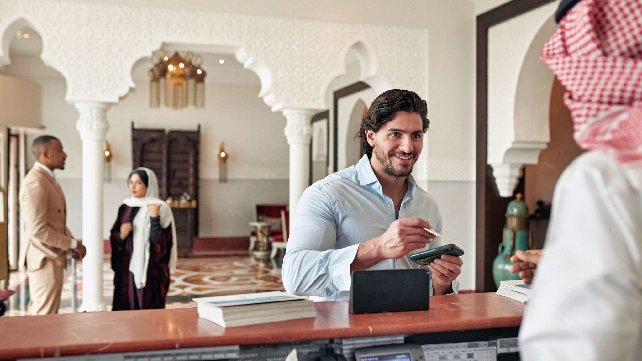Ein Mann checkt an einer Hotelrezeption bei einem Mitarbeiter mit rot-weiß kariertem Kopftuch ein in einer Lobby mit orientalischer Bauweise.