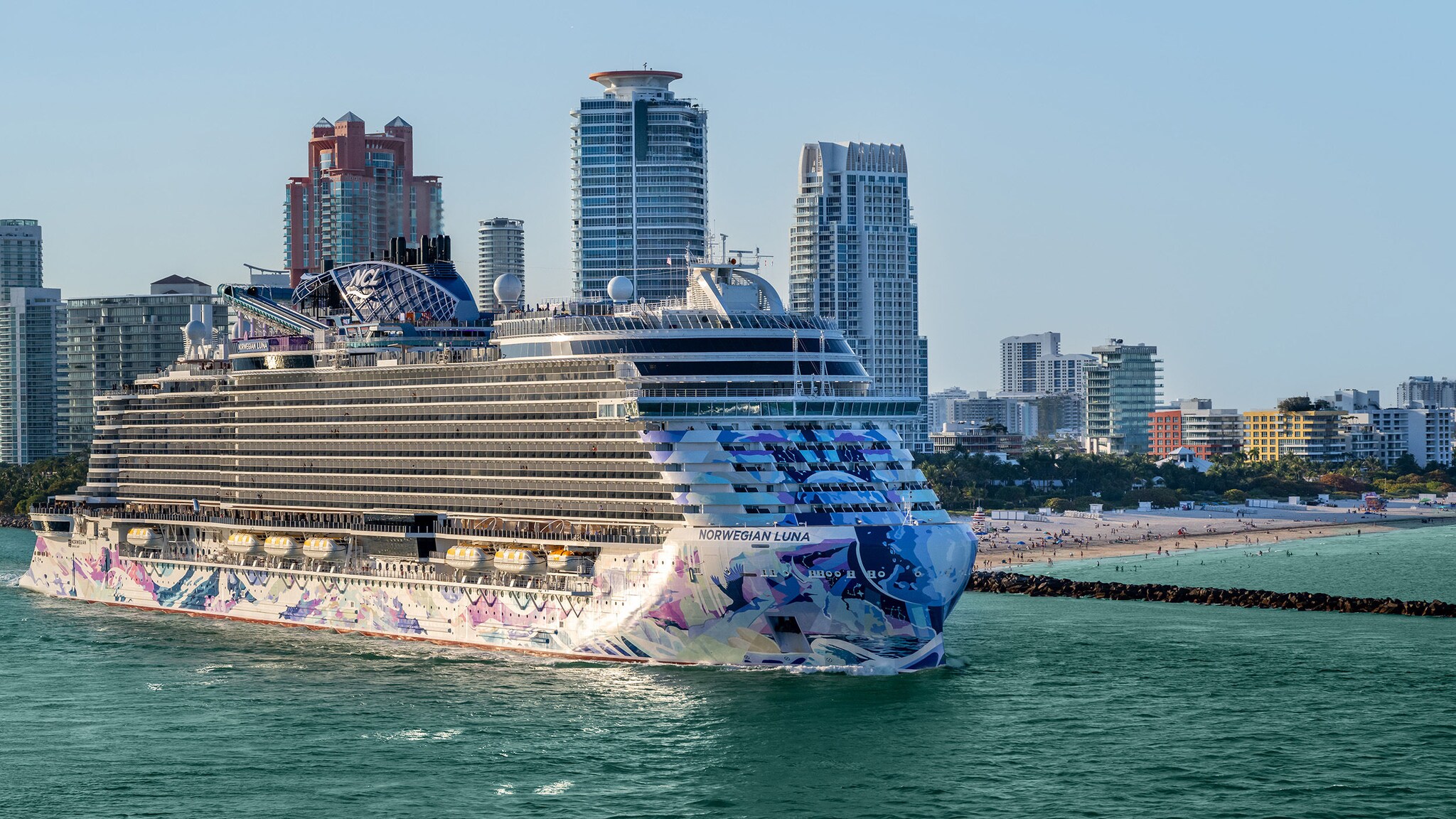 Ein Kreuzfahrtschiff mit Bordwand im bunten Design am Strand vor der Skyline von Miami.