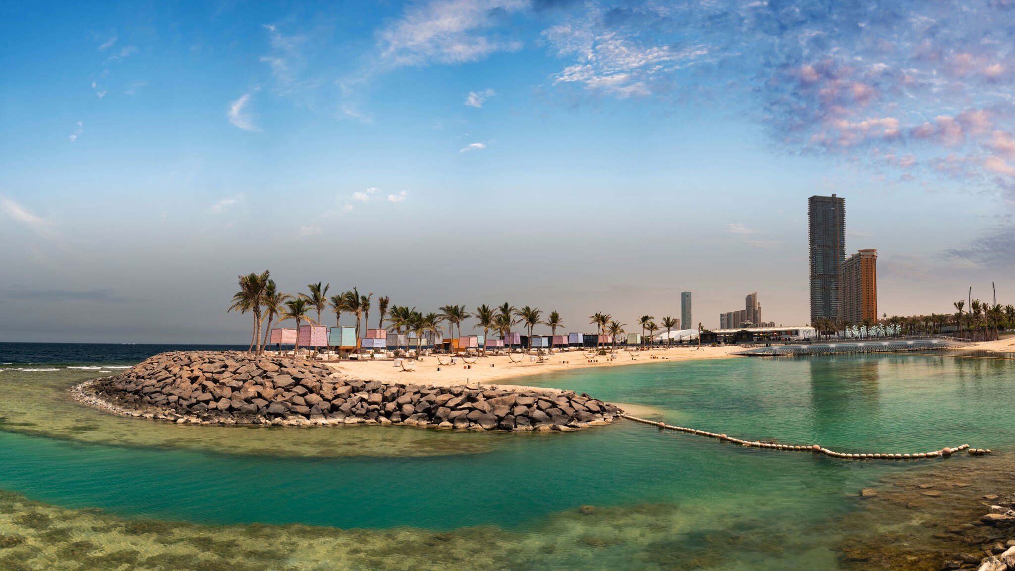 Landzunge mit Palmen und bunten Bungalows an einem hellen Sandstrand, umgeben von türkisgrünem Wasser, im Hintergrund Hochhäuser.