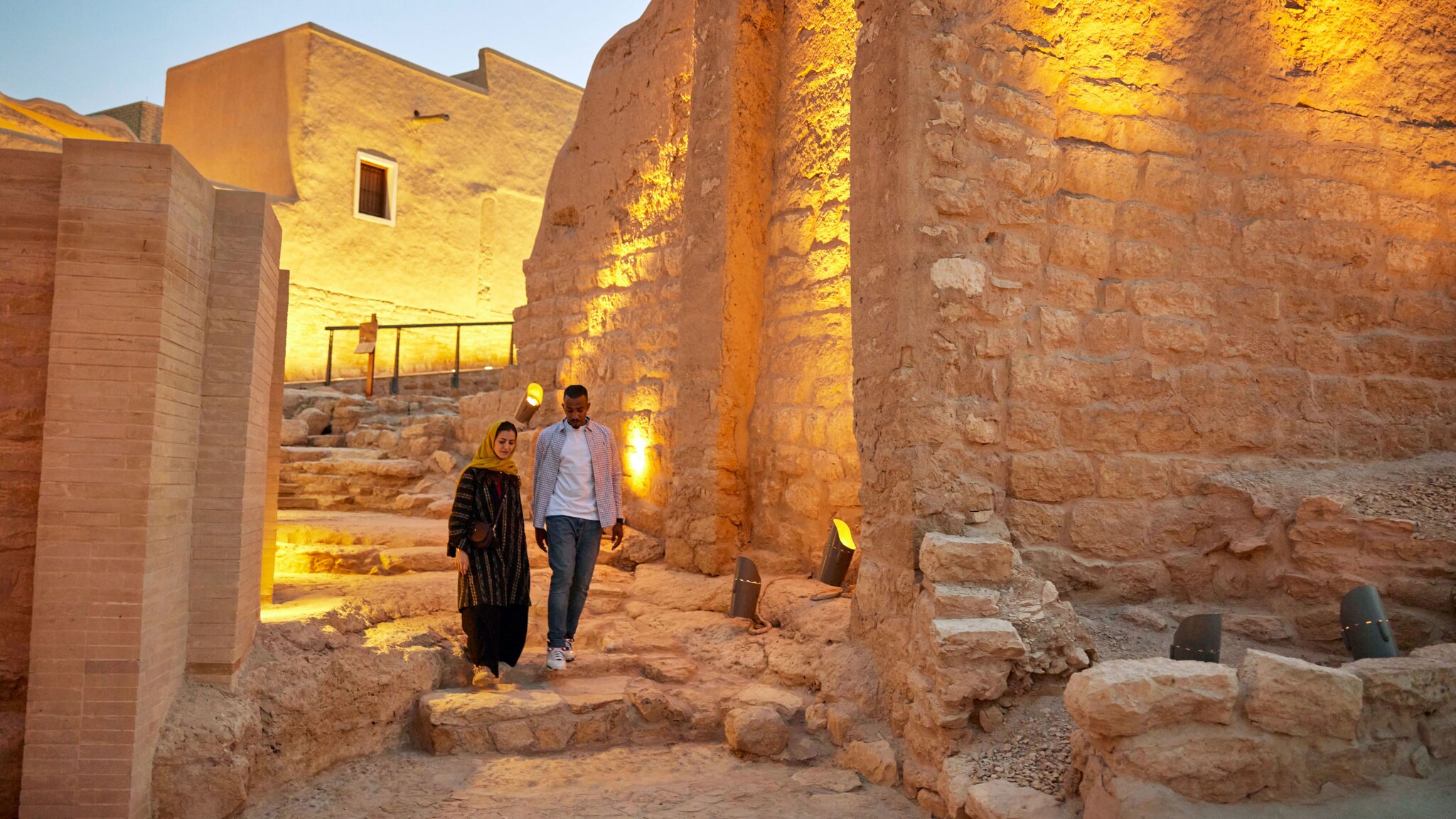 Ein Paar läuft über eine Steintreppe durch eine historische Stätte mit angeleuchteten Lehmbauten.