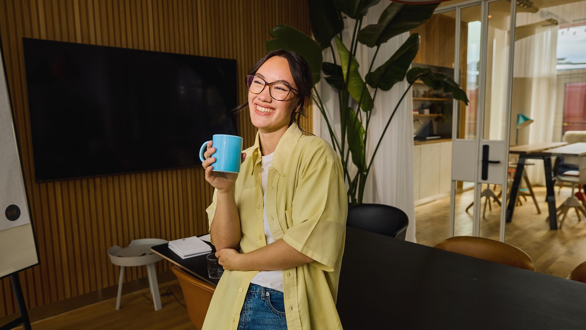 Eine lächelnde Frau lehnt entspannt mit Kaffeetasse an einem Schreibtisch in einem Büro.