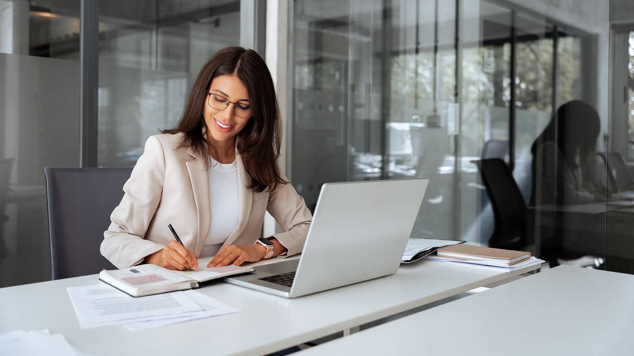 Eine Frau im weißen Blazer arbeitet an einem Schreibtisch mit Dokumenten und Laptop in einem Büro.