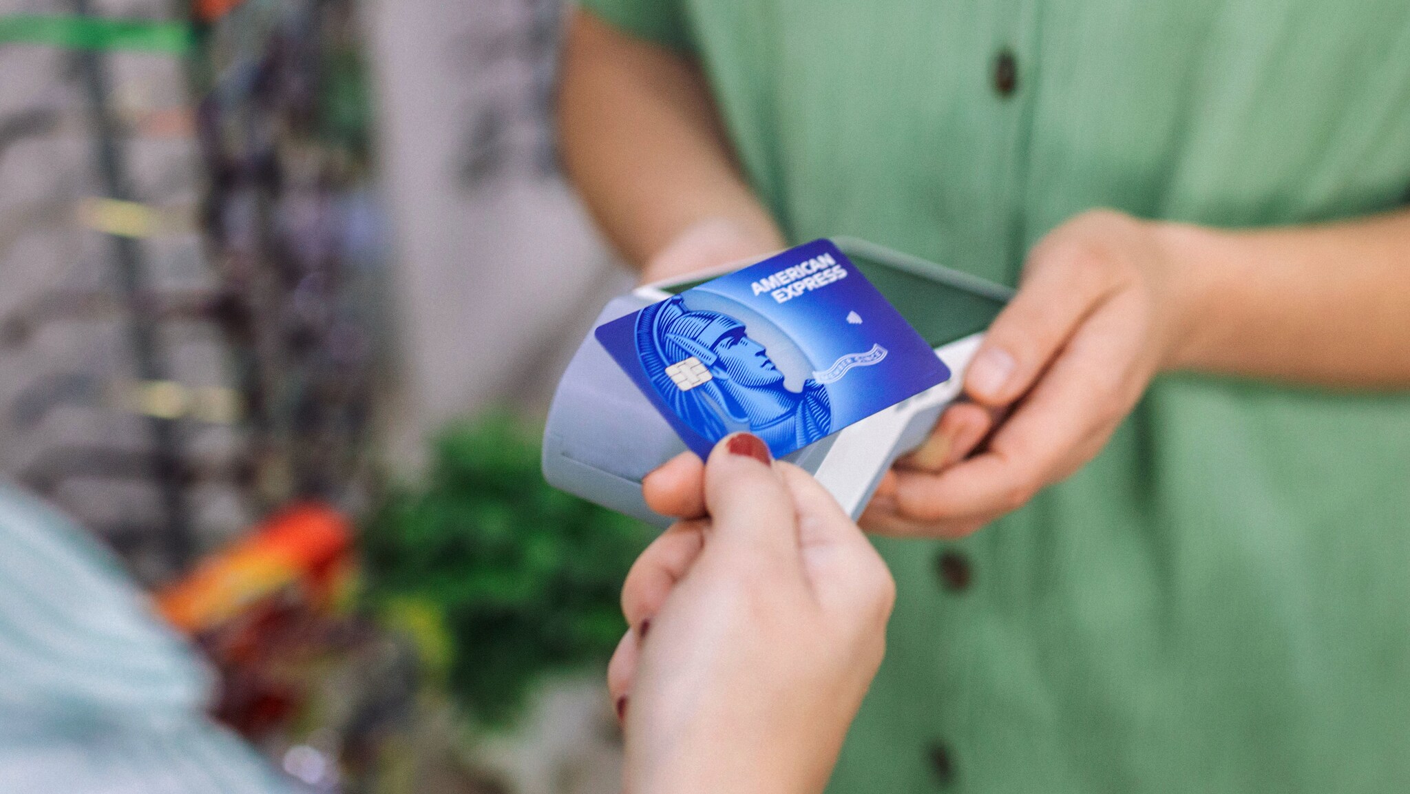 Nahaufnahme einer kontaktlosen Zahlung mit einer blauen Kreditkarte von American Express an einem Kartenlesegerät in der Hand einer Person in einem Geschäft.
