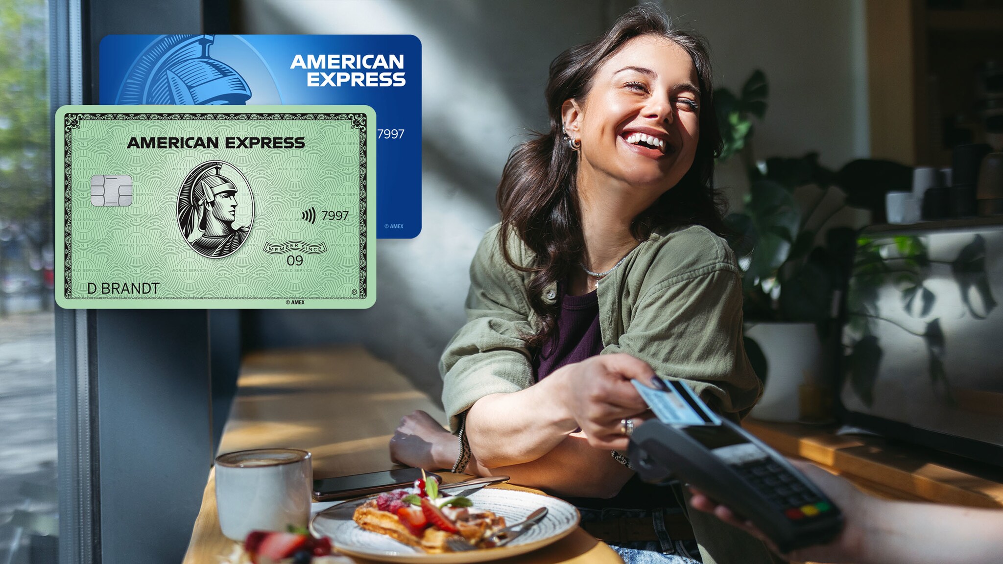 Eine junge Frau sitzt am Fenster eines Cafés und zahlt gut gelaunt mit einer Kreditkarte kontaktlos an einem Kartenlesegerät, im Vordergrund eine grüne und eine blaue Kreditkarte von American Express als Fotomontage.