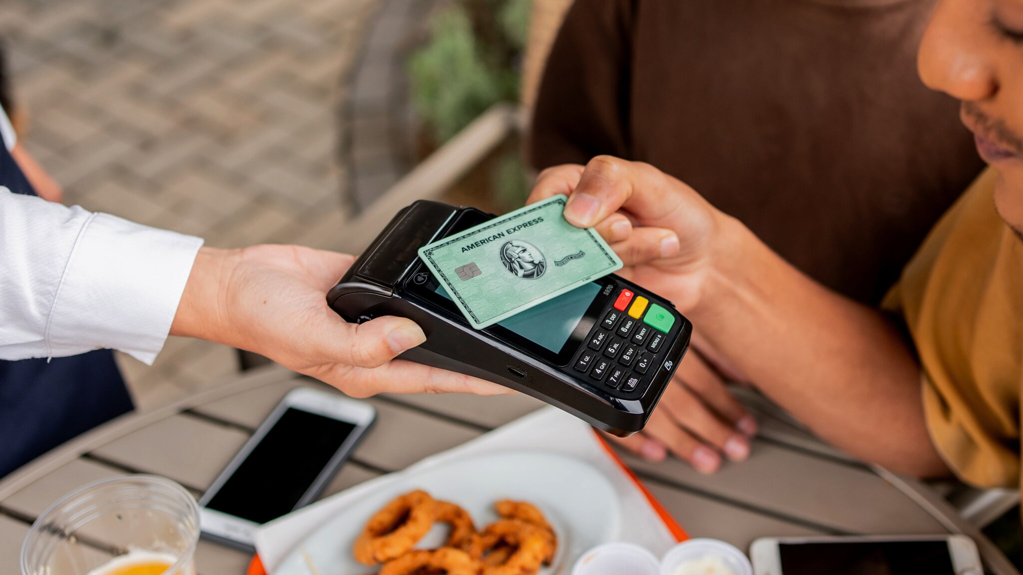 Nahaufnahme einer kontaktlosen Zahlung mit einer grünen Kreditkarte von American Express an einem Kartenlesegerät in der Hand einer Person in einem Straßencafé.