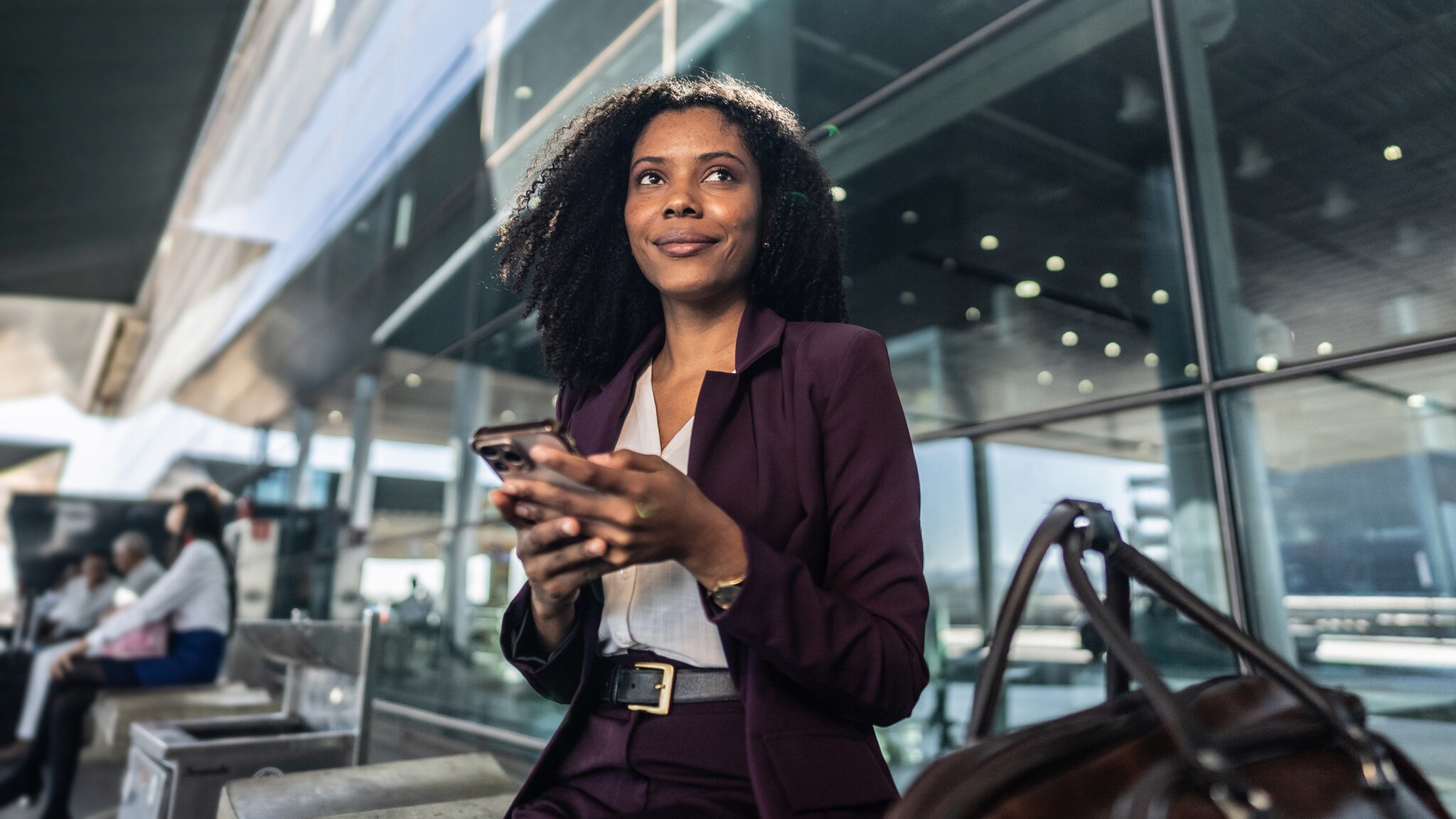 amex-sbs-amex-platinum-business-versicherungen-vergleich-flughafenterminal Eine elegant gekleidete Business-Frau sitzt mit einem Smartphone in ihren Händen neben einer Reisetasche in einem Flughafenterminal und blickt lächelnd in den Raum.