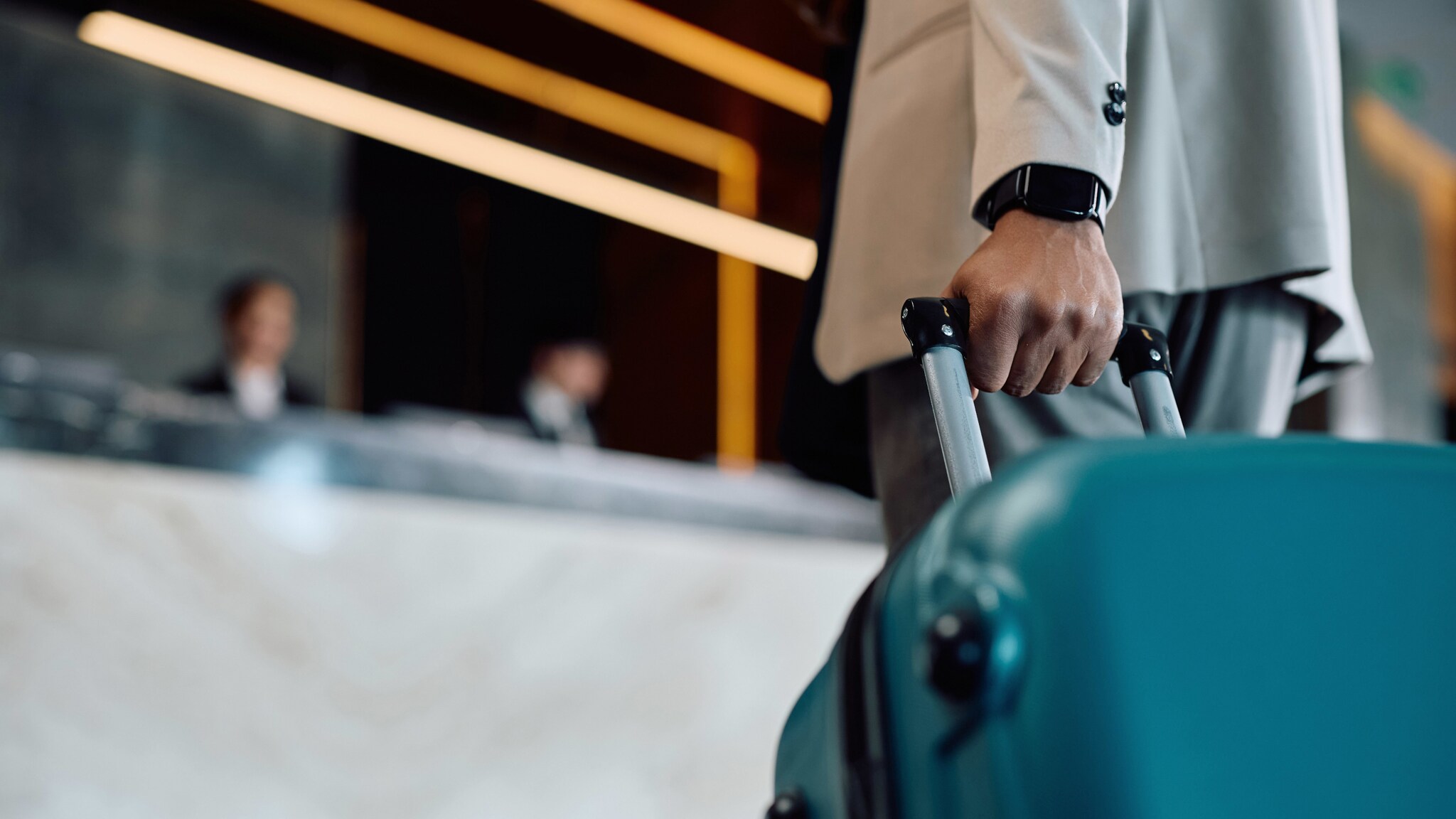 Die Hand eines elegant gekleideten Mannes am Griff eines türkisfarbenen Hartschalenkoffers vor einer Hotelrezeption.