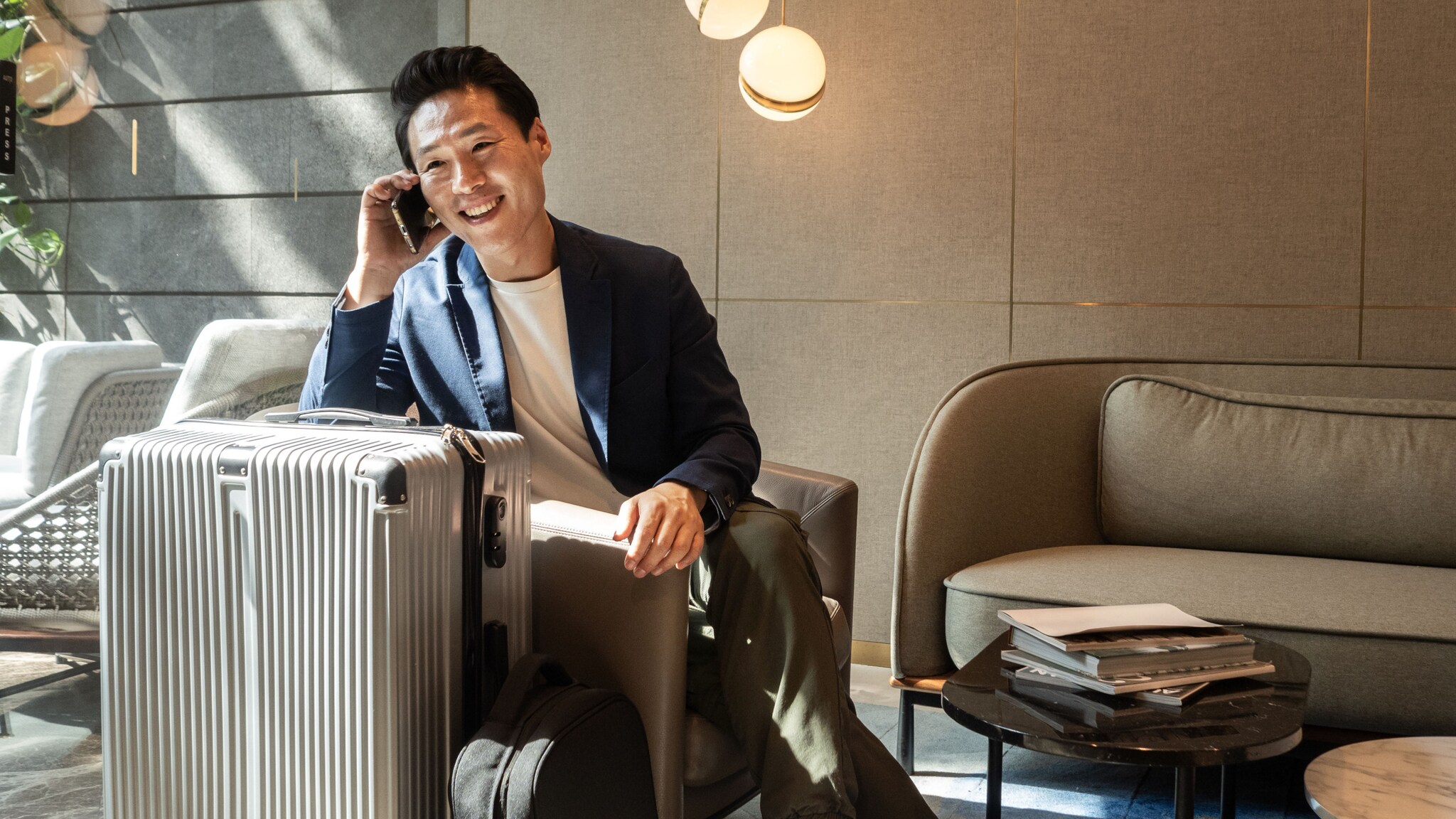 Ein Business-Mann sitzt neben einem großen Reisekoffer in einem Sessel in einer Lounge und hält lächelnd ein Smartphone an sein Ohr.