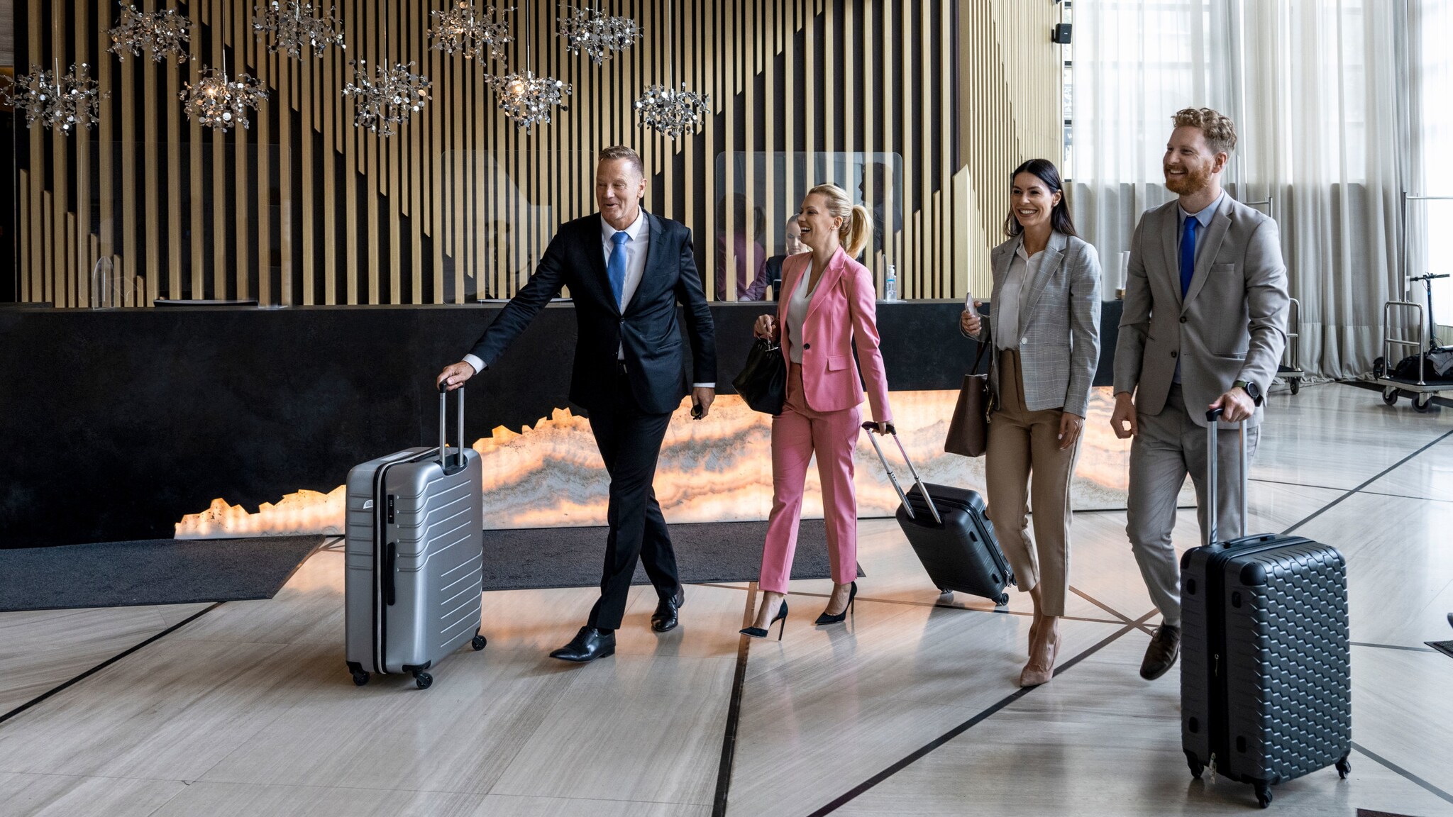 Vier Personen in Business-Kleidung gehen mit Reisegepäck nebeneinander in einer modernen Hotellobby.