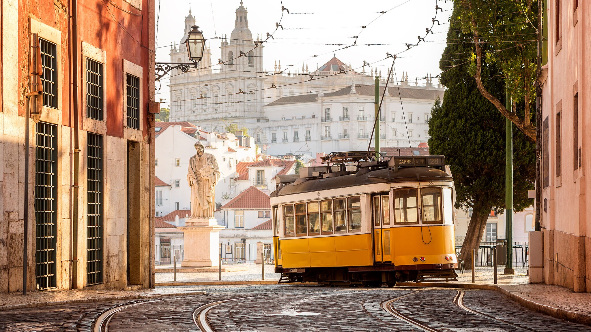 Ein gelbe historischer Straßenbahn in der Altsatdt von Lissabon neben einer Statue an einem Platz vor einer weißen Kathedrale. 