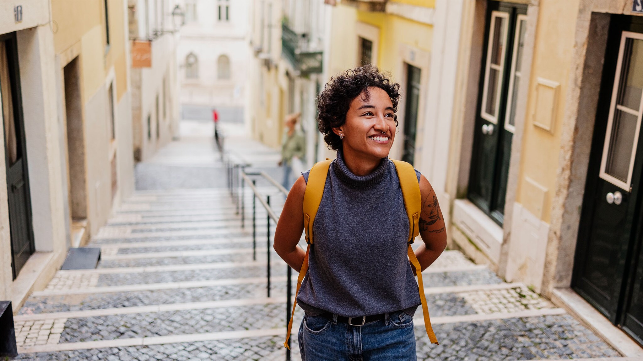Eine junge Frau mit Rucksack läuft lächelnd eine Treppe in einer Gasse in einer mediterranen Altstadt hinauf.