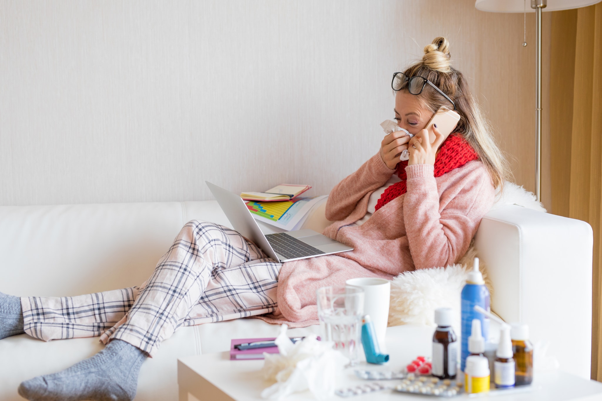 amex-insurance-kann-ich-meine-reise-wegen-krankheit-stornieren-frau-laptop Eine Frau sitzt mit einem Laptop auf dem Sofa, sie telefoniert und schnäuzt sich die Nase, vor ihr Medikamente.