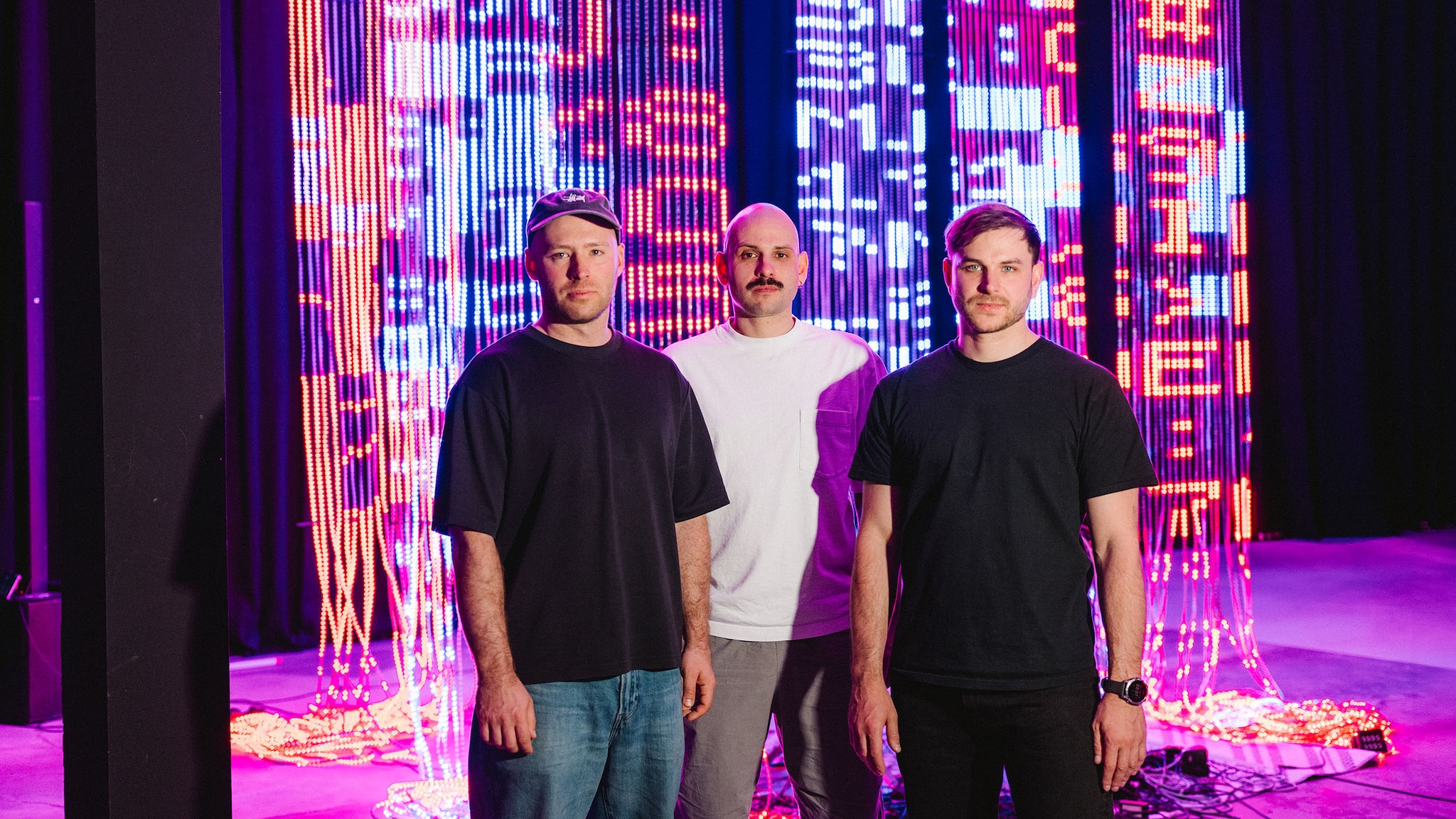 amex-magazin-amex-art-on-ksawery-komputery-drei-maenner Drei Männer posieren vor einer hängenden Lichtinstallation aus leuchtenden LED-Streifen in einer Halle.