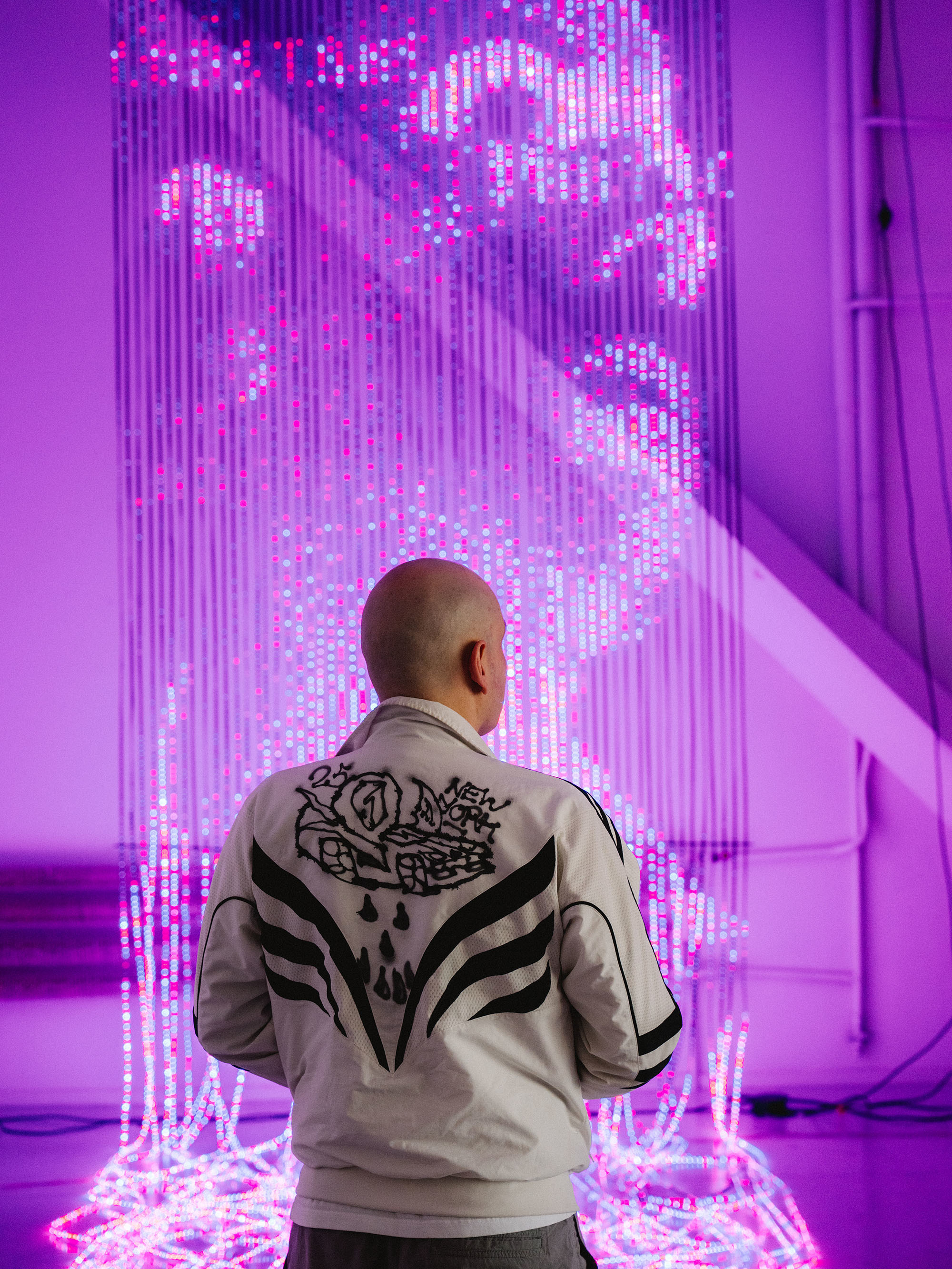 Rückansicht eines Mannes, der vor einer hängenden Lichtinstallation mit einem Gesichtsmotiv aus pink leuchtenden LEDs steht.