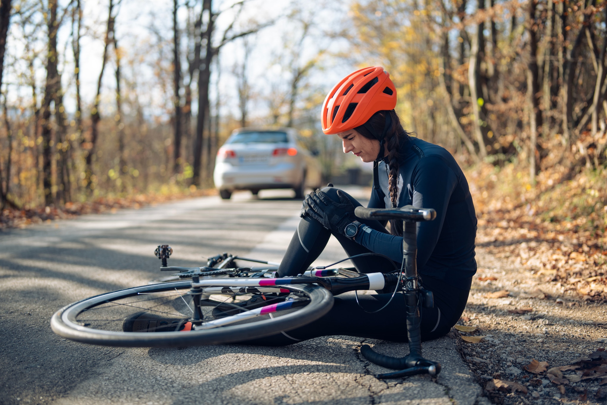 Eine Frau mit Fahrradhelm sitzt neben ihrem Rennrad auf der Straße und hält sich das Knie, im Hintergrund steht ein Auto.