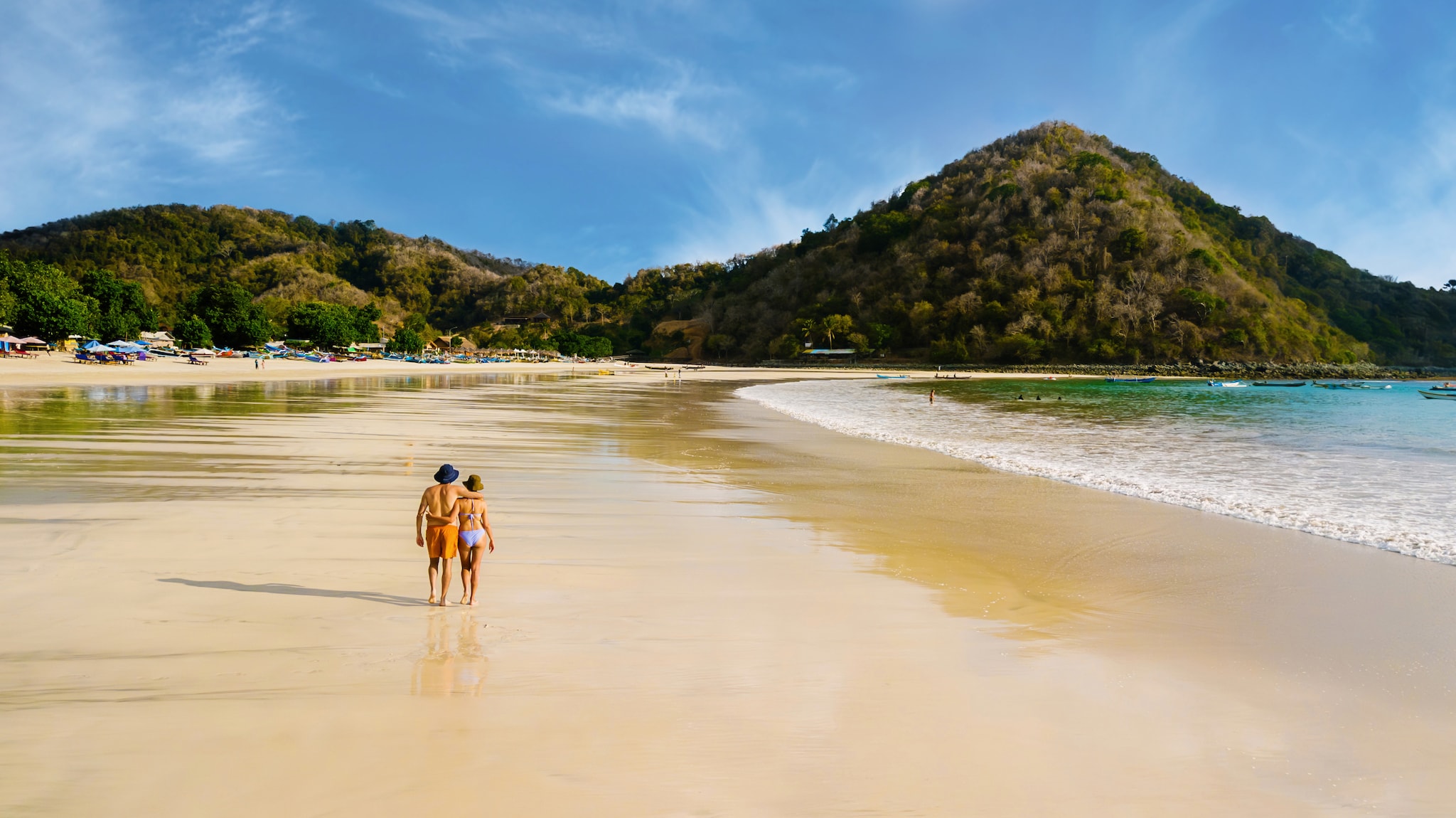 Ein Mann und eine Frau spazieren am breiten Sandstrand entlang, im Hintergrund grün bewachsene Berge.