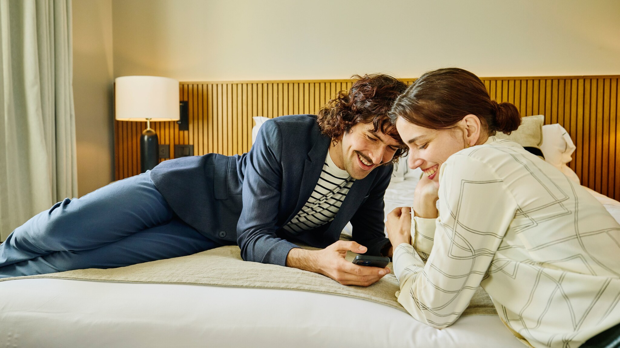 Ein Mann und eine Frau liegen quer auf einem Bett und schauen lächelnd gemeinsam auf ein Smartphone in seiner Hand.