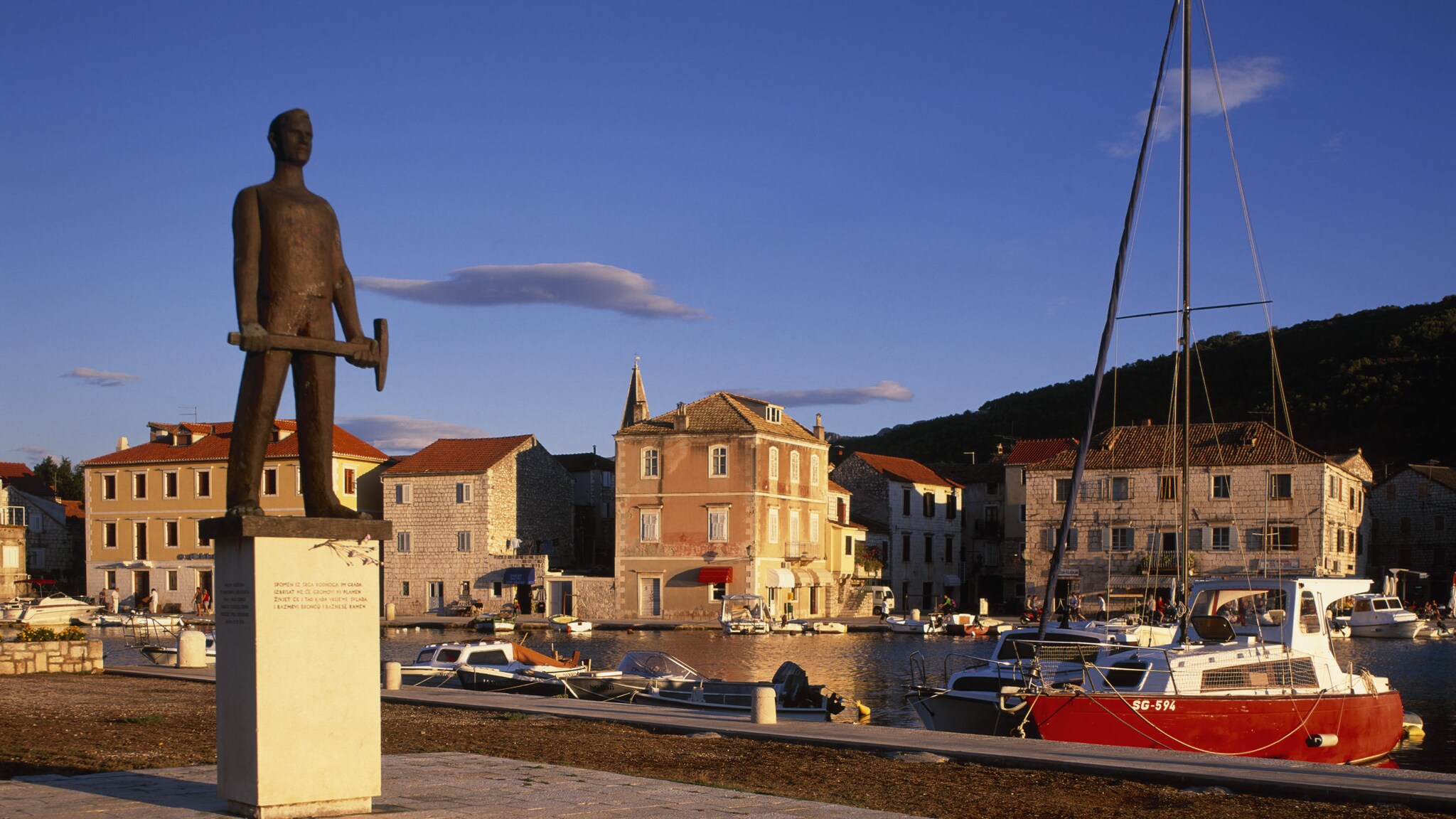 Blick auf Häuser und Statue des kleinen Hafenorts Stari Grad mit kleinen Booten im Wasser.