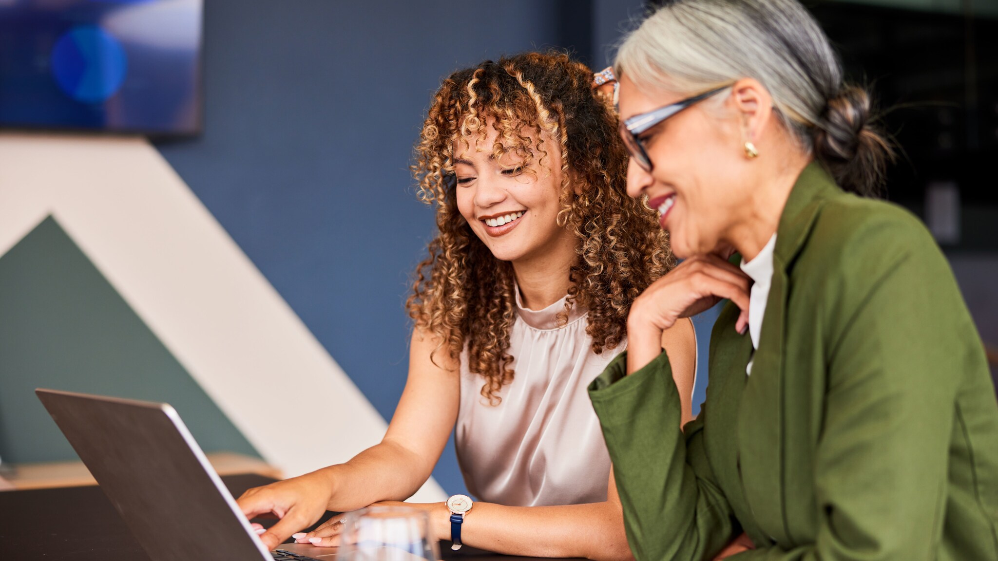Eine junge Frau mit Locken und eine ältere Frau mit grauen Haaren sitzen gut gelaunt nebeneinander und blicken gemeinsam auf einen aufgeklappten Laptop in einem Konferenzraum.