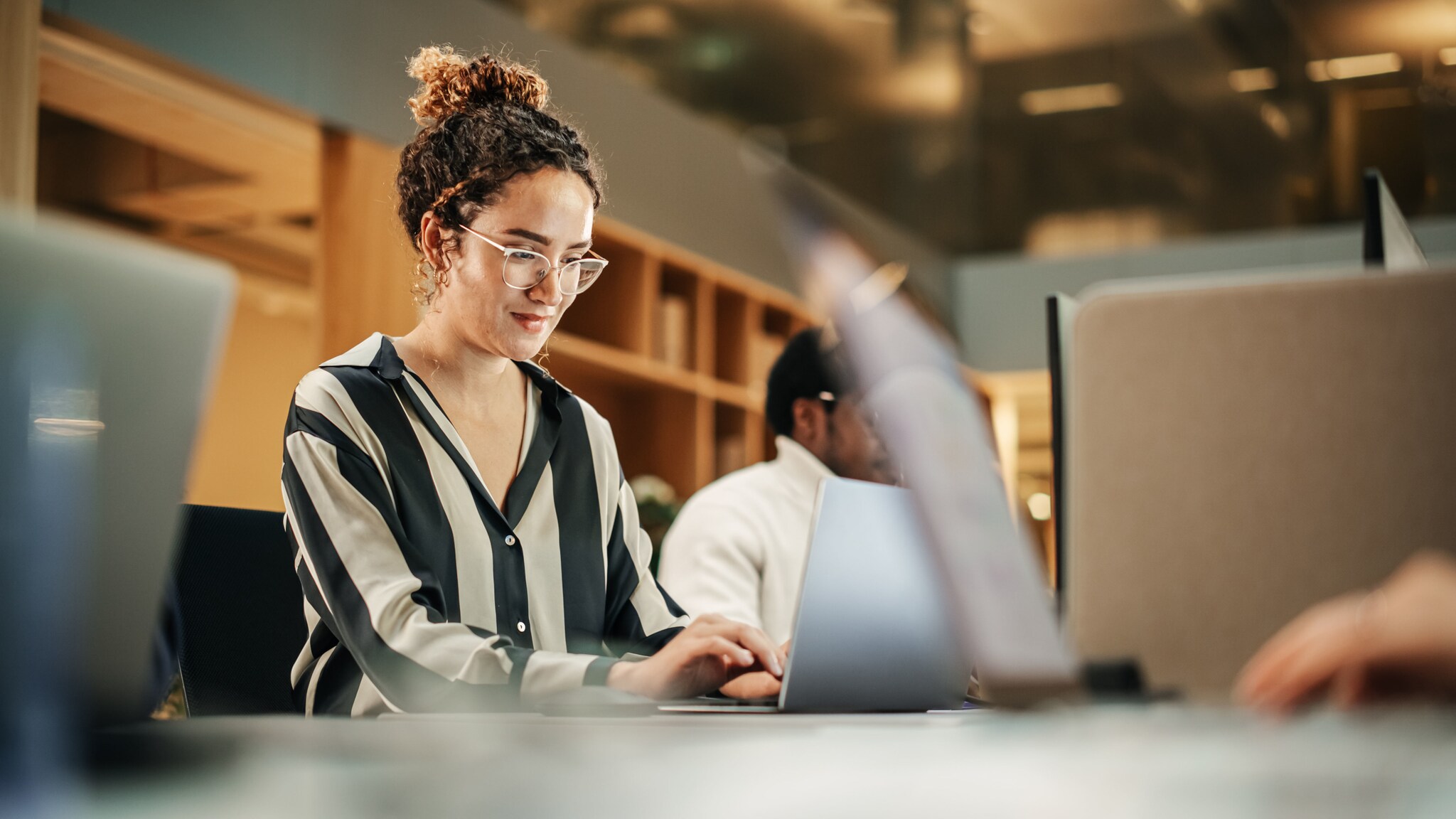 Eine modern gekleidete Frau arbeitet an einem Laptop an einem Tisch mit weiteren Personen in einem Büro.