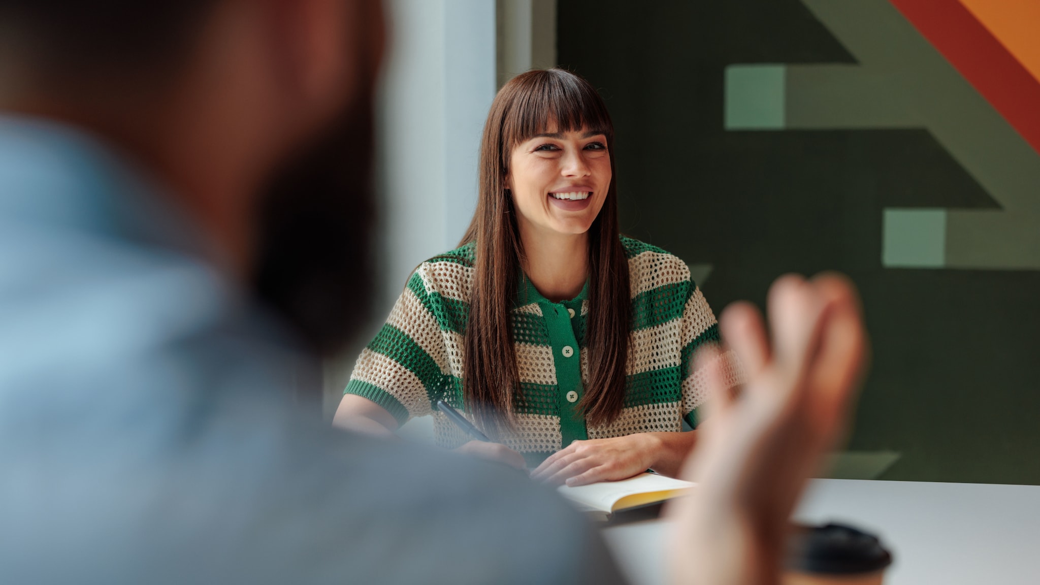 amex-sbs-arbeitsplatzwechsel-innerhalb-des-unternehmens-frau Eine junge Frau mit Ponyfrisur sitzt mit Notizblock an einem Tisch vor einer gemusterten Wand und schaut lächelnd zu einer ihr gegenübersitzenden Person.