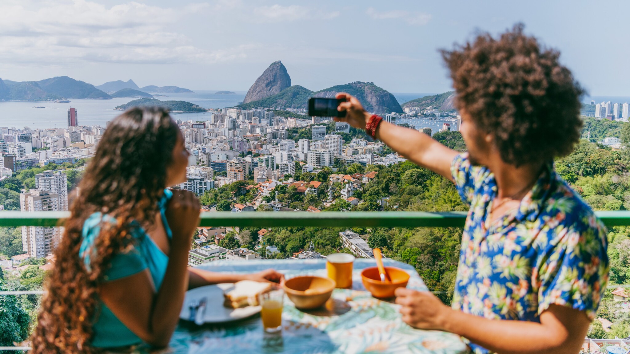 Profilansicht von einer Frau und einem Mann, die sich gegenüber an einem Tisch mit Frühstück sitzen auf einer Terrasse im Sonnenschein über der Skyline von Rio de Janeiro am Meer, er hält ein Smartphone zum Fotografieren hoch.