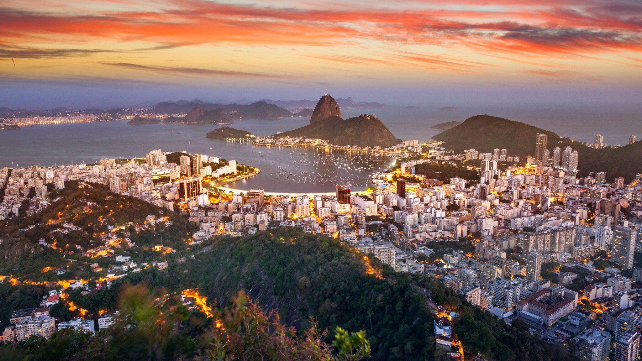 Stadtpanorama von Rio de Janeiro, umgeben von grüner Hügellandschaft am Meer bei Sonnenuntergang aus der Luftperspektive.