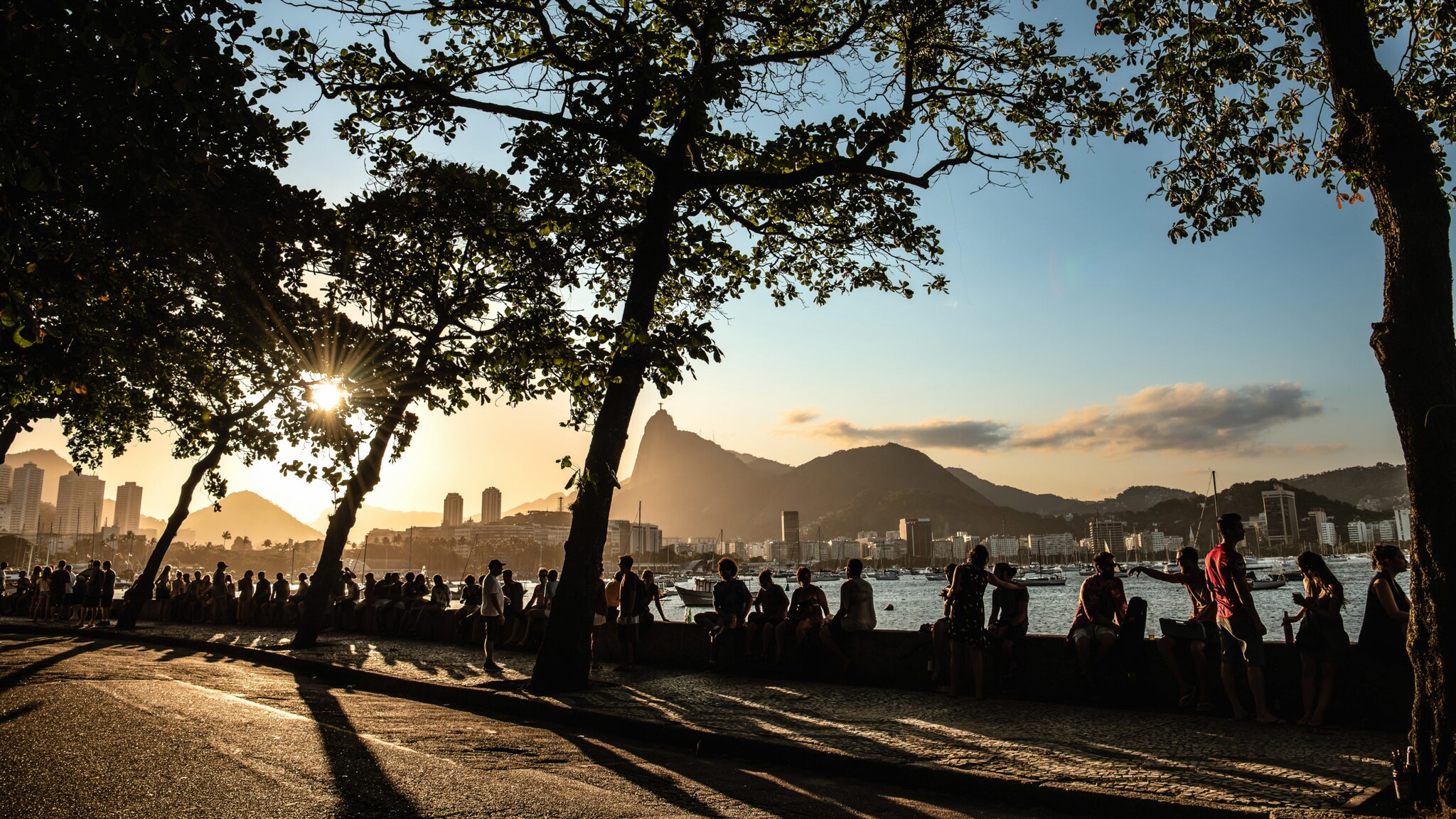 Silhouetten zahlreicher sitzender Personen auf einer Mauer an einer Bucht mit Stadtpanorama im Gegenlicht bei Sonnenuntergang.