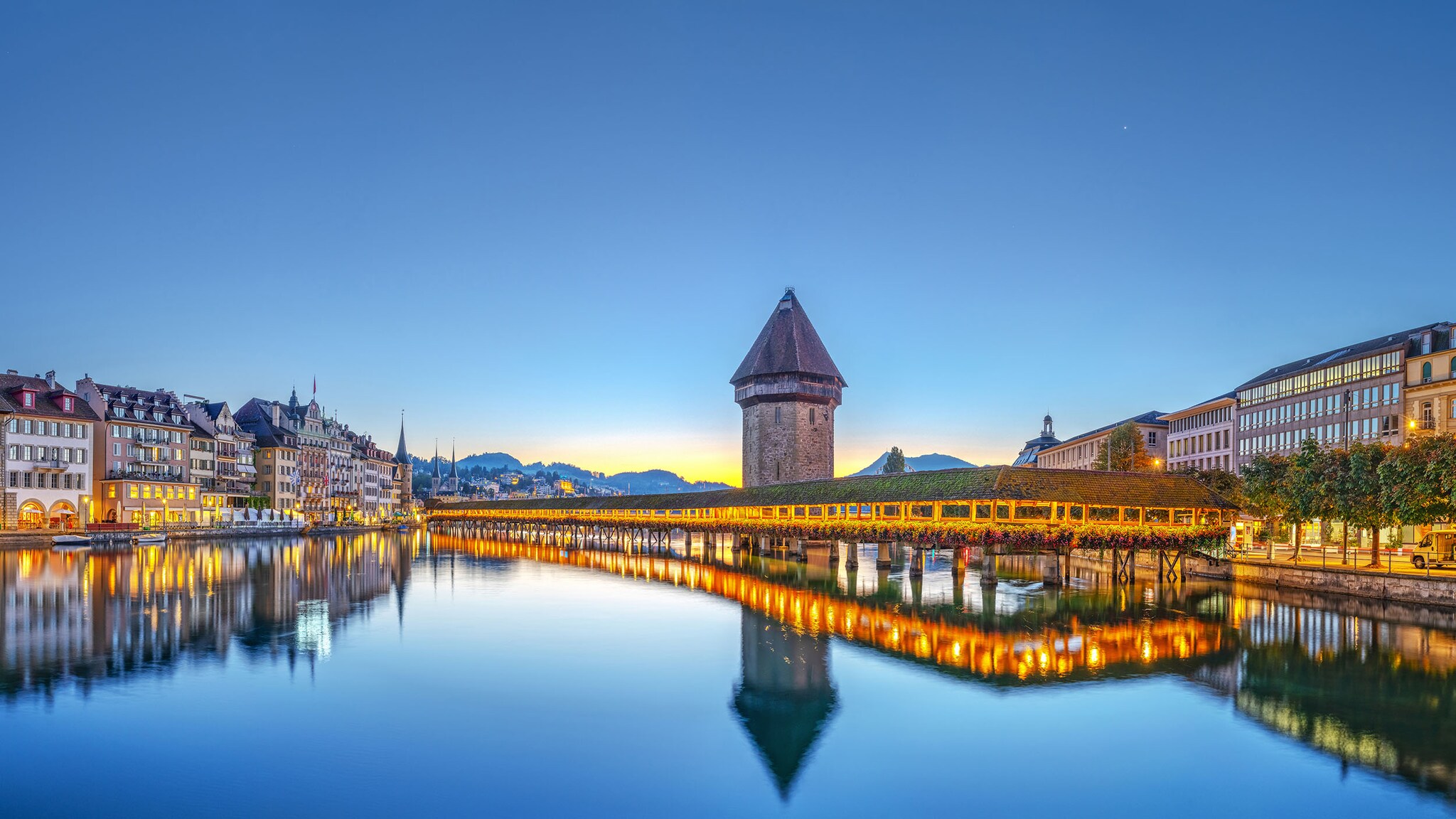 Über einen Fluss führt eine historische Holzbrücke, am Ufer stehen historische Gebäude, die beleuchtet sind.