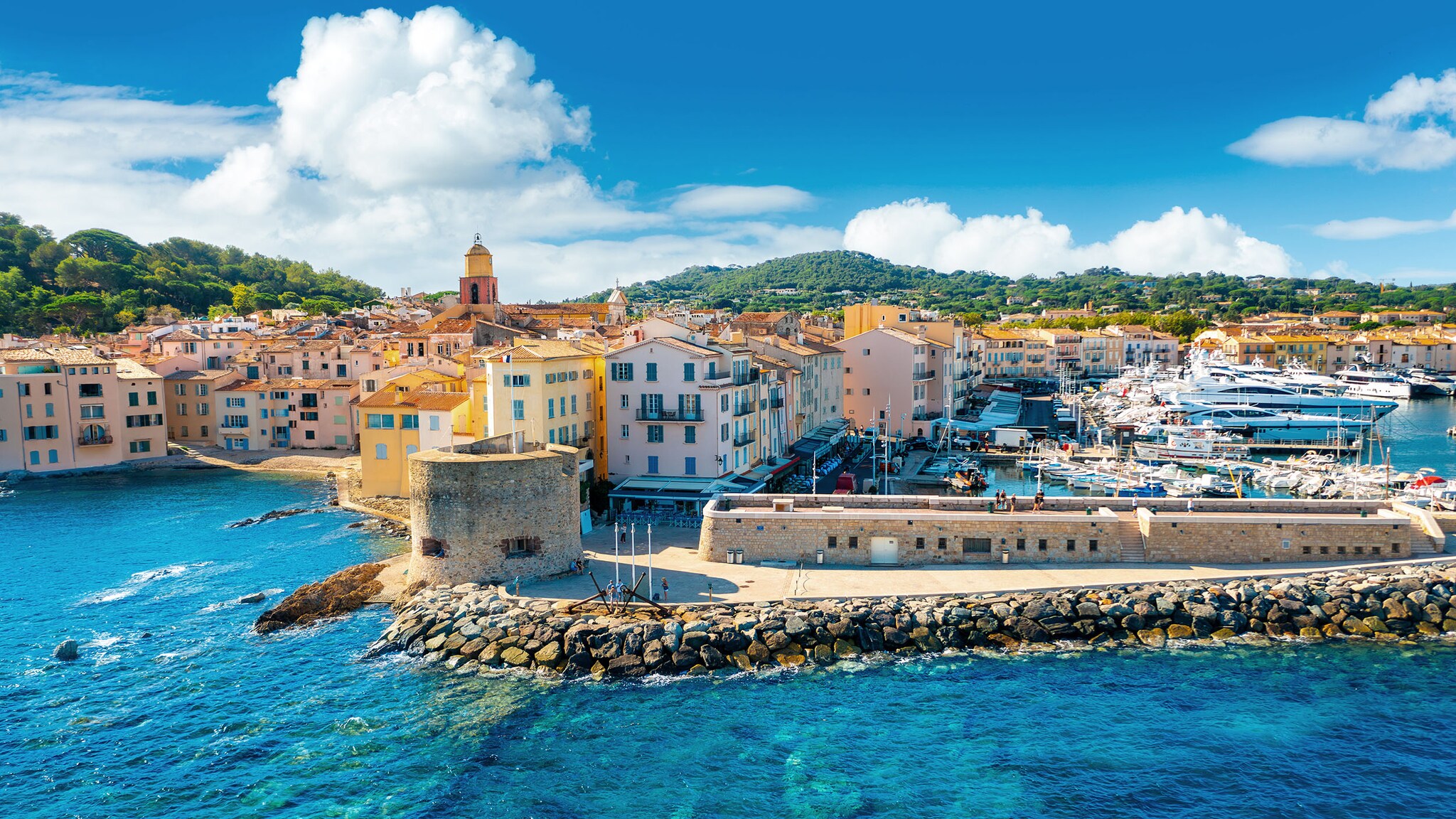 Panorama-Ansicht von Saint-Tropez mit Mole und pastellfarbenen Häuserzeilen der Altstadt und Hafenbecken mit zahlreichen Booten im azurblauen Wasser bei Sonnenschein.