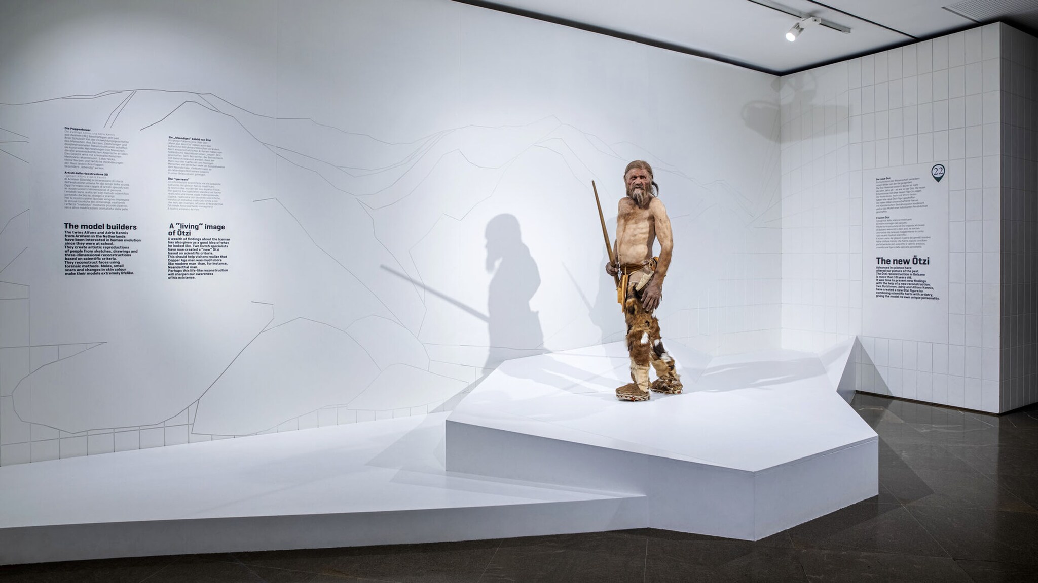 Aufrechtstehendes Ötzi-Modell in Kleidung aus Fell und Leder mit Speer in der Hand auf einem weißen Podest in einem Ausstellungsraum.
