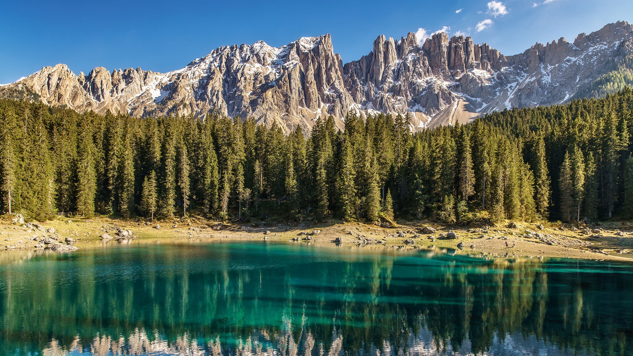 Klarer, grün schimmernder Bergsee, umgeben von Fichtenwald, vor zerklüftetem Bergpanorama mit Schneeflecken unter blauem Himmel bei Sonnenschein.