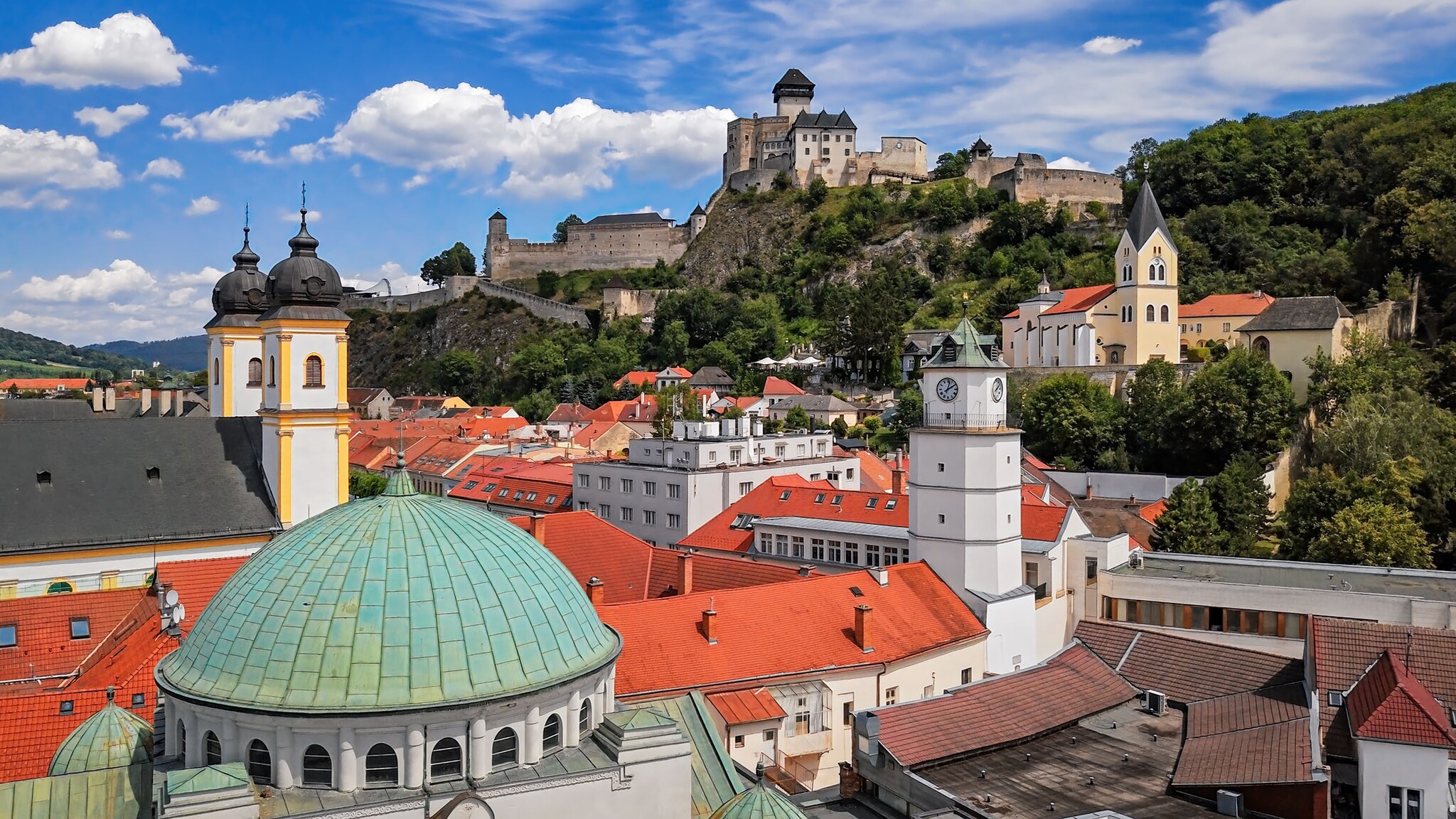 Historische Altstadt mit Kirchtürmen vor einem bewaldeten Berg mit Burg unter blauem Himmel.